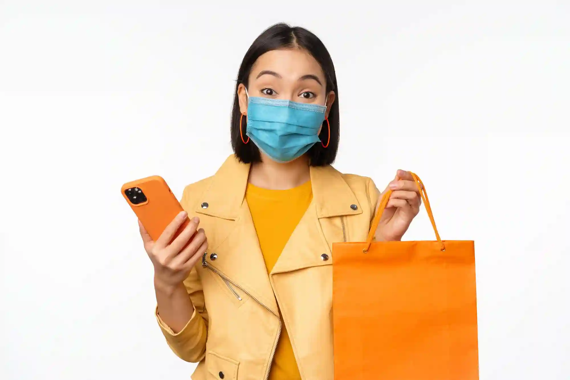 A masked woman holding an orange shopping bag and smartphone, highlighting the rise of safe mobile-enabled retail experiences.