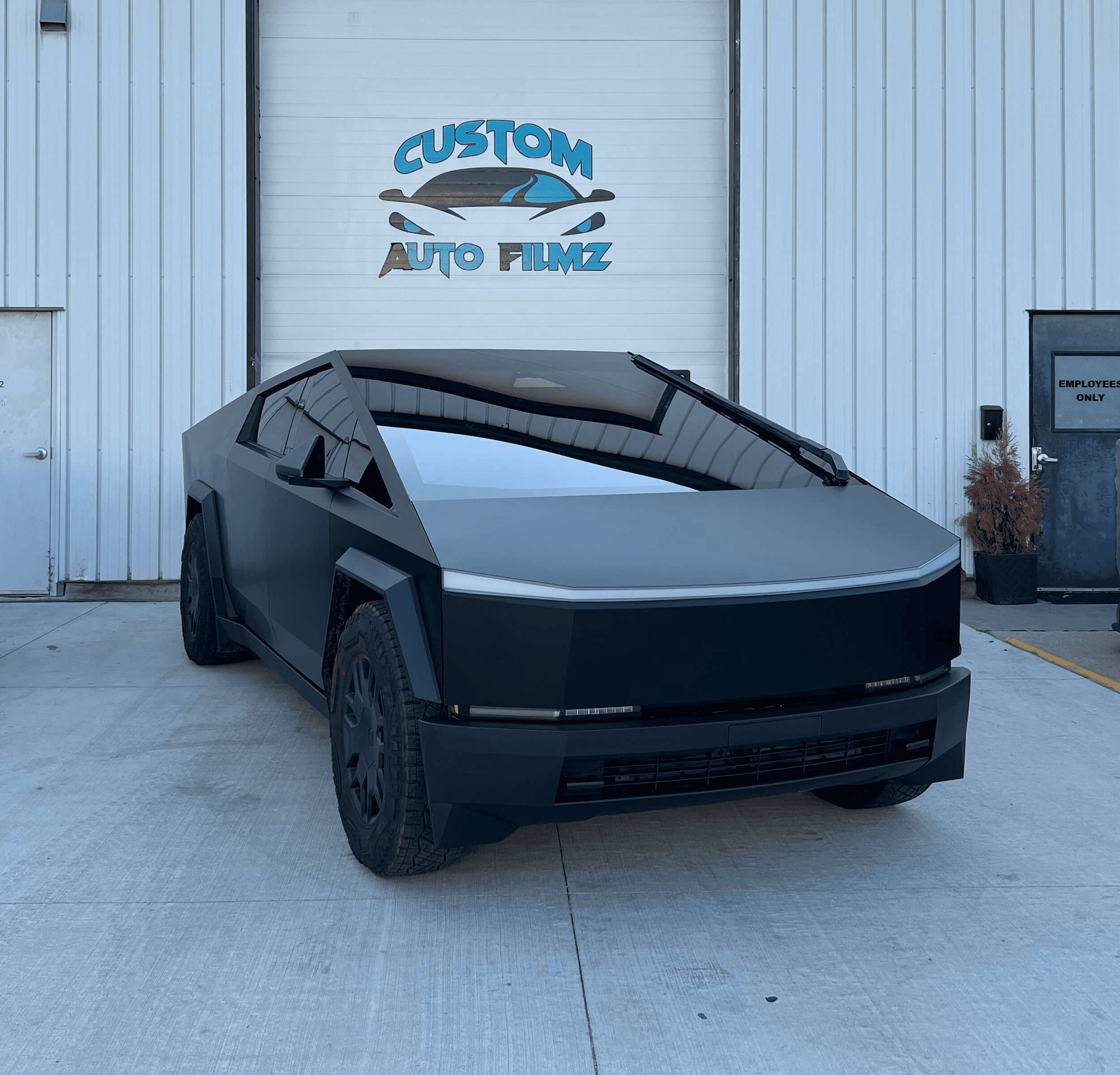 A matte black Tesla Cybertruck is parked on a concrete driveway in front of a white industrial building.