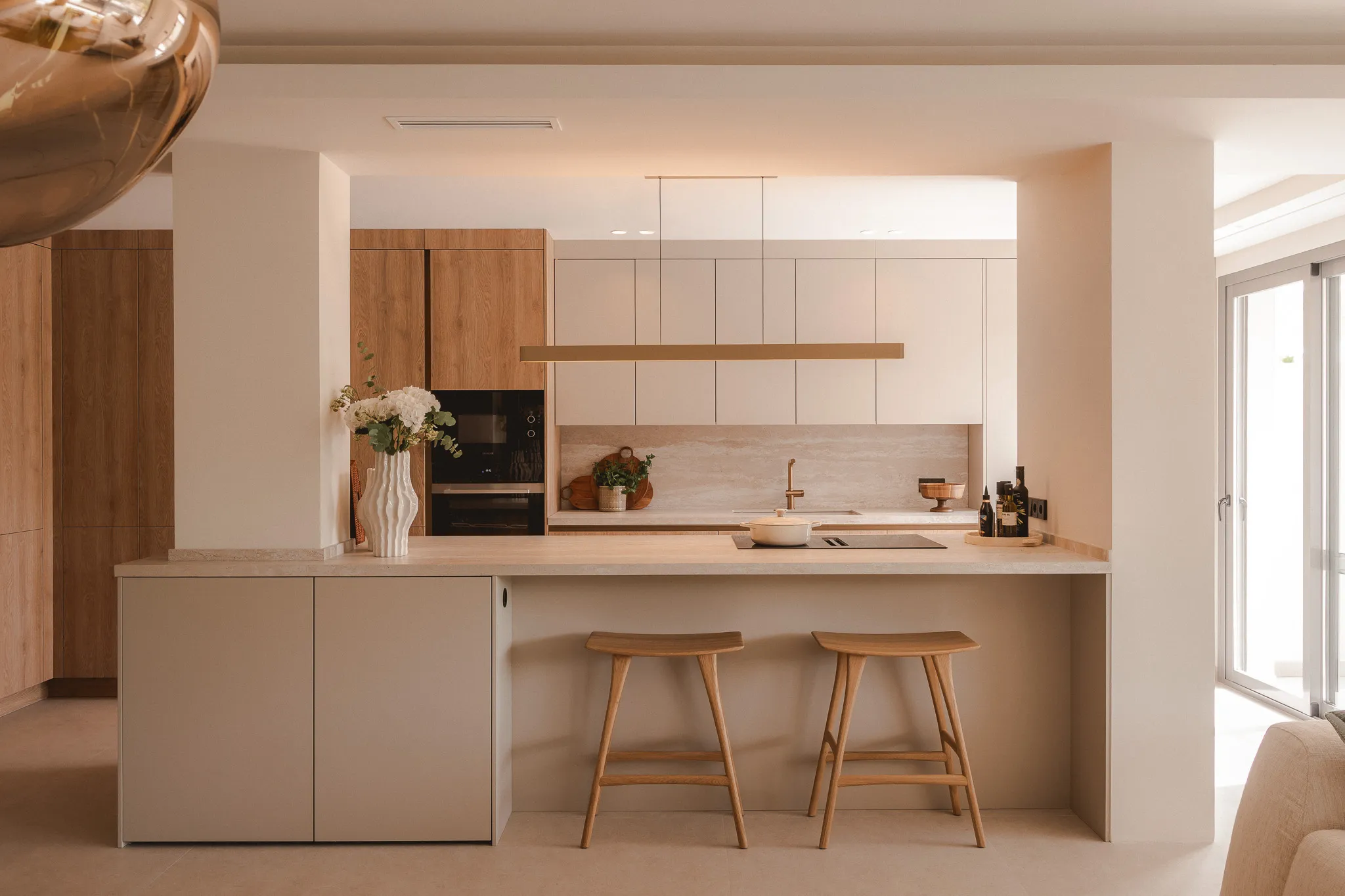 A modern, minimalist kitchen with a large beige island, two light wood bar stools, and warm wood cabinetry in the background.