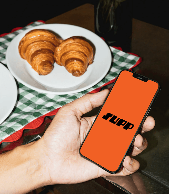 A hand holding a phone displaying the Supp app, with croissants on a cafe table in the background