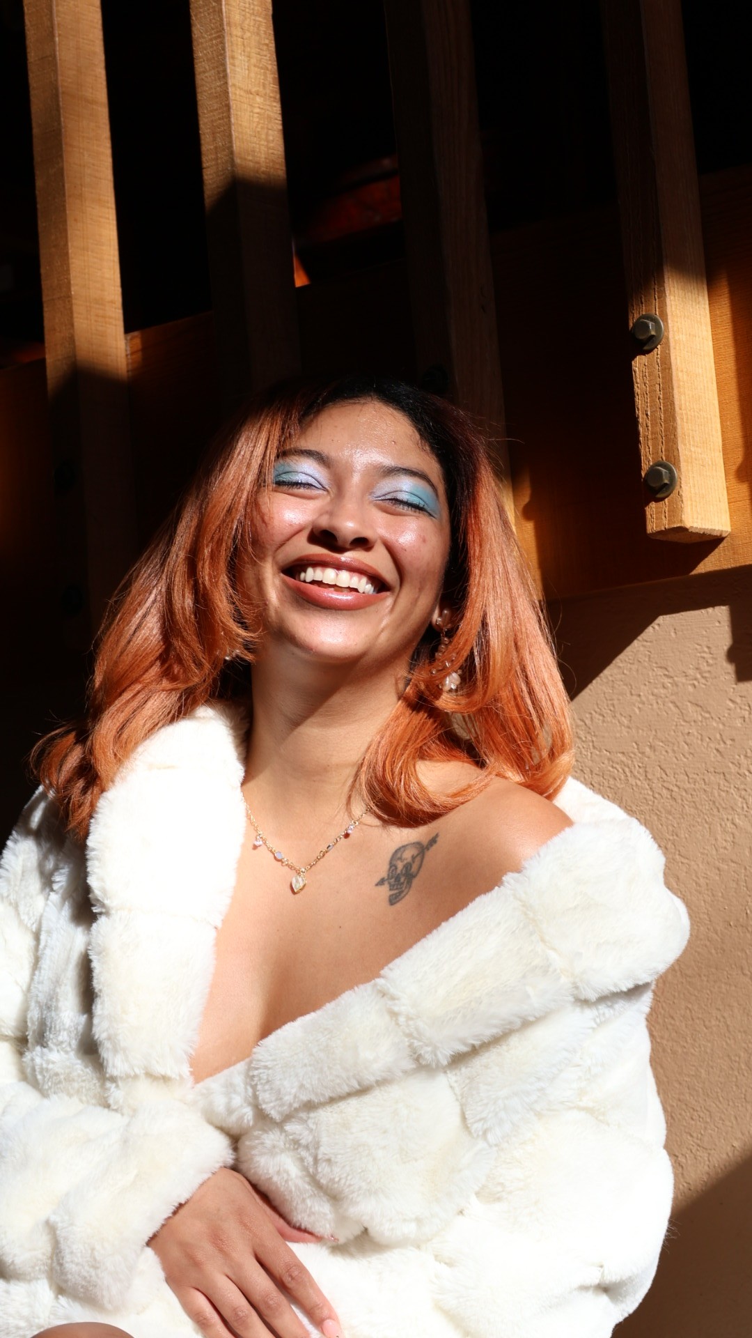 A brown woman with red hair and a white fur coat on closing her eyes and smiling