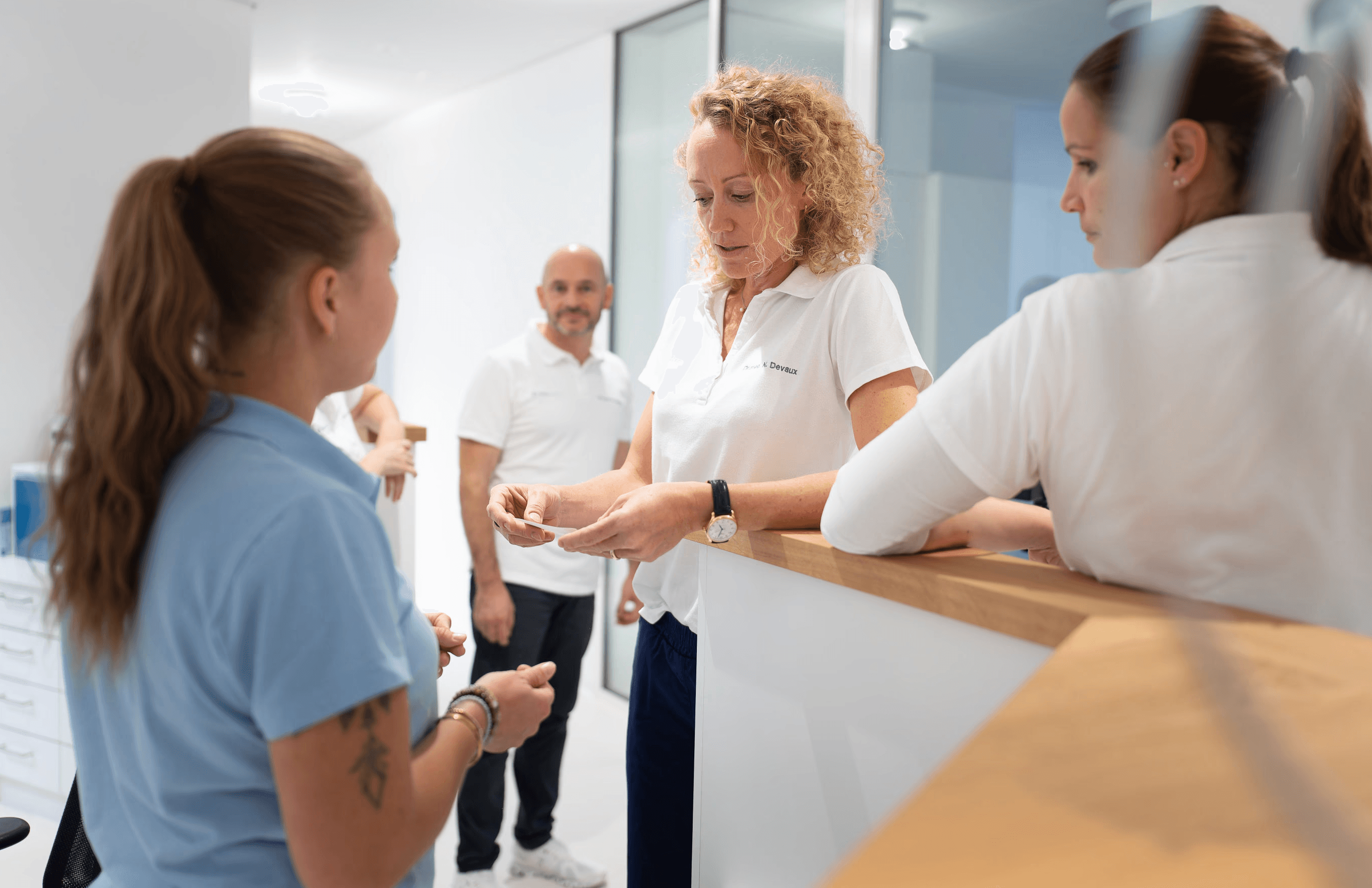 Vier Personen in der modernen, hellen Handklinik unterhalten sich miteinander. Dr. Noémie Devaux prüft etwas in ihren Händen, während zwei Mitarbeitende zuschauen. Alle tragen professionelle weisse oder hellblaue Hemden.