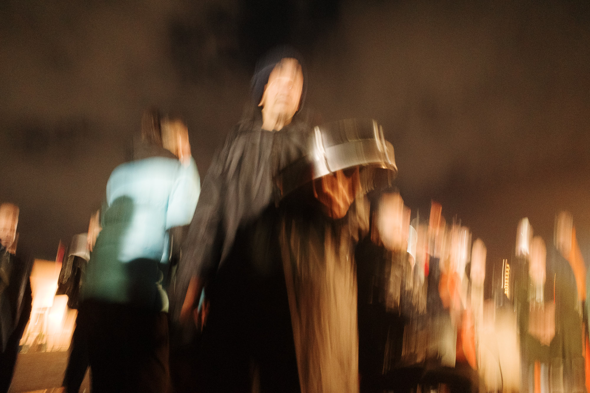 Blurred image of people gathered outdoors at night, with a person holding a reflective object under a partly cloudy sky.
