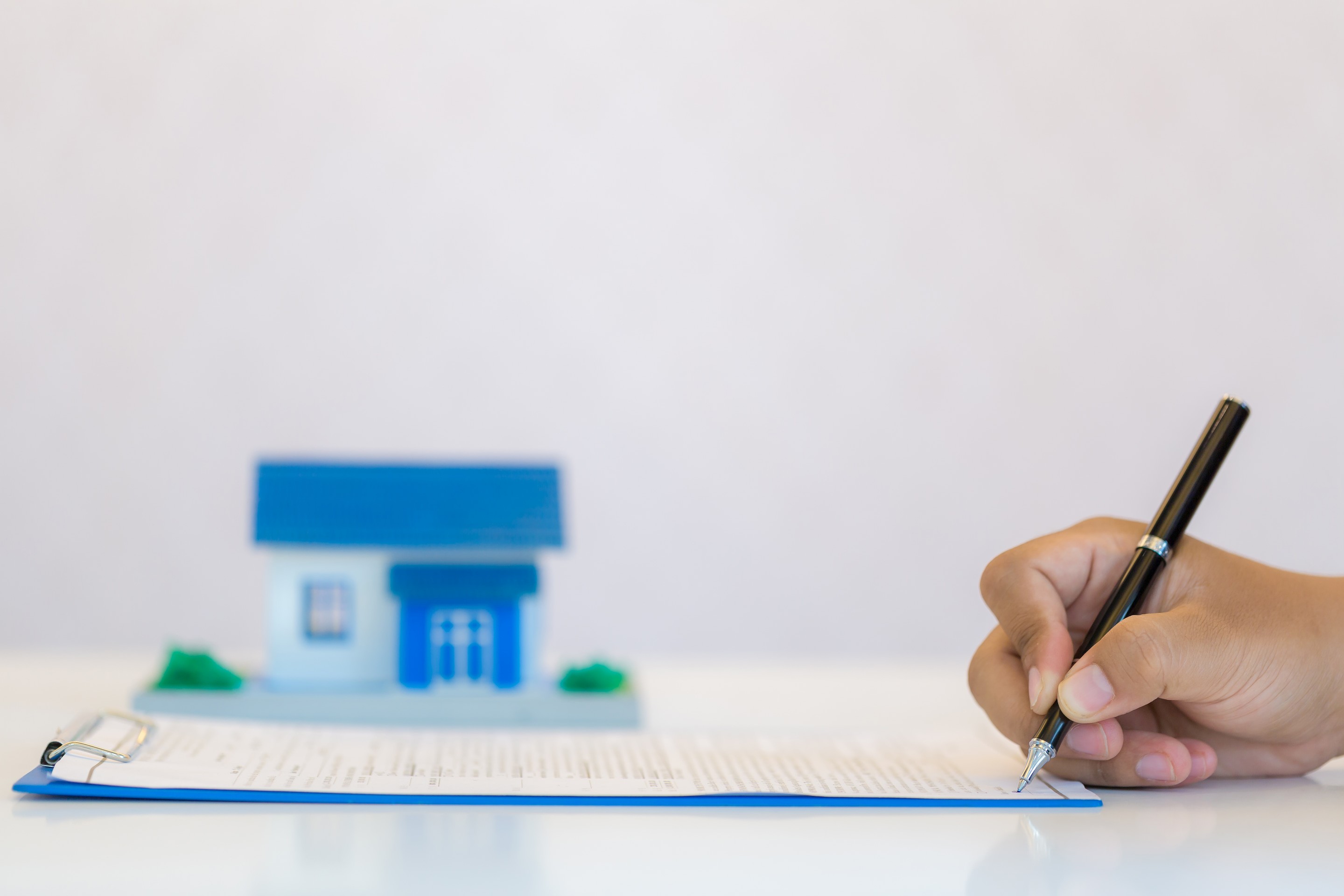 A hand signing a document on a clipboard with a small model house in the background.