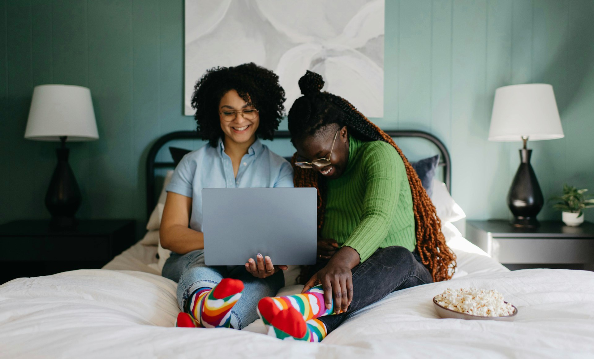 Two women, one in a light blue shirt and the other in a green sweater, are sitting together on a bed, looking down and laughing at a laptop resting on their laps.