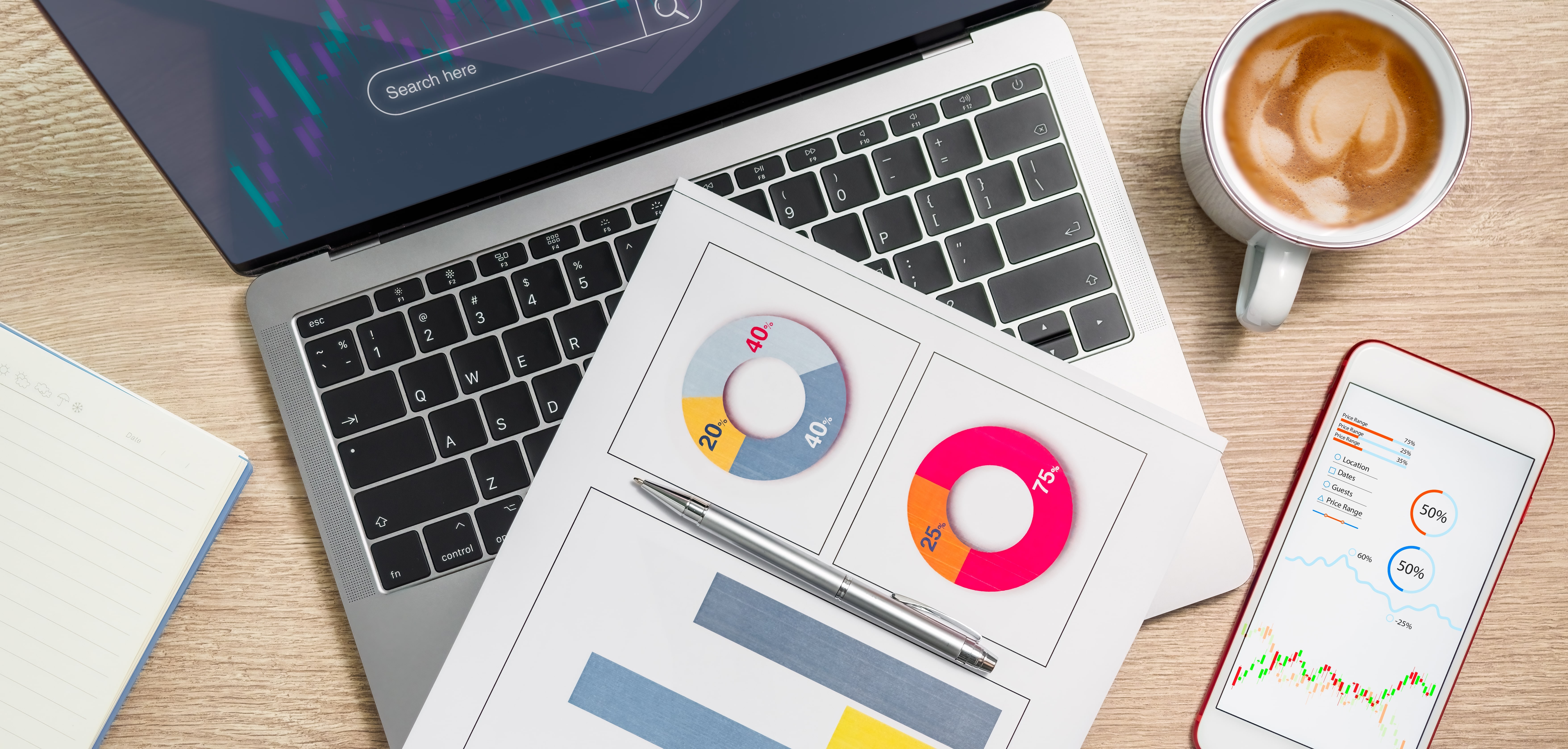 An overhead view of a modern desk prepared for data analysis, featuring a laptop and smartphone displaying financial dashboards and performance charts. Printed business reports with colorful pie and bar graphs are arranged next to a cup of coffee, illustrating market research and financial planning.