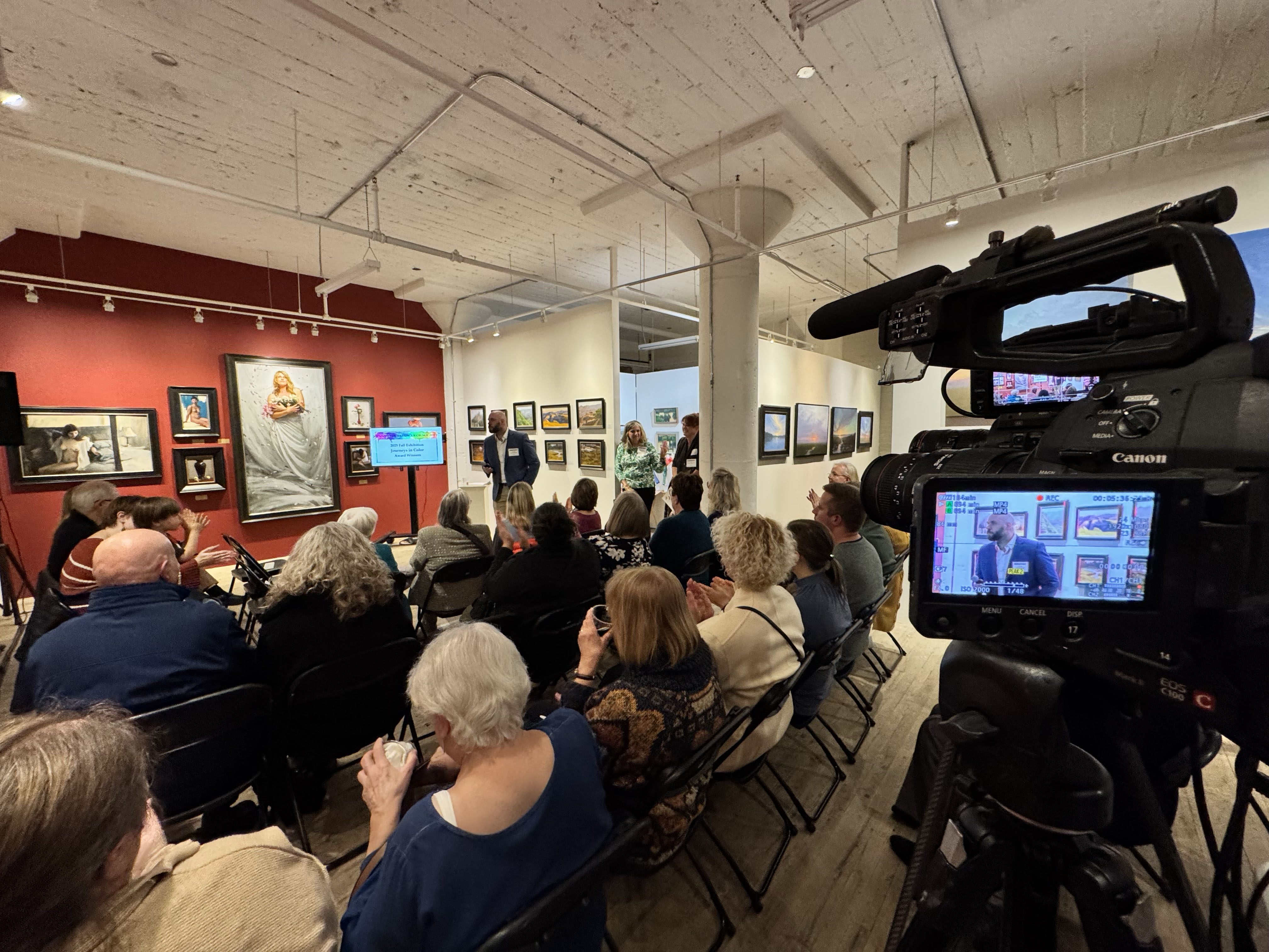A video camera records a live presentation and audience inside an art gallery with framed artwork on display.
