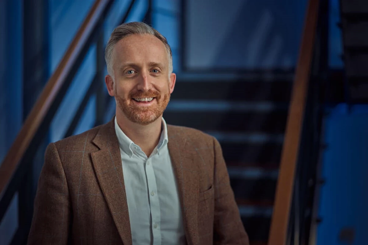 A person wearing a brown blazer and white shirt stands indoors in front of a staircase, with soft blue lighting in the background.
