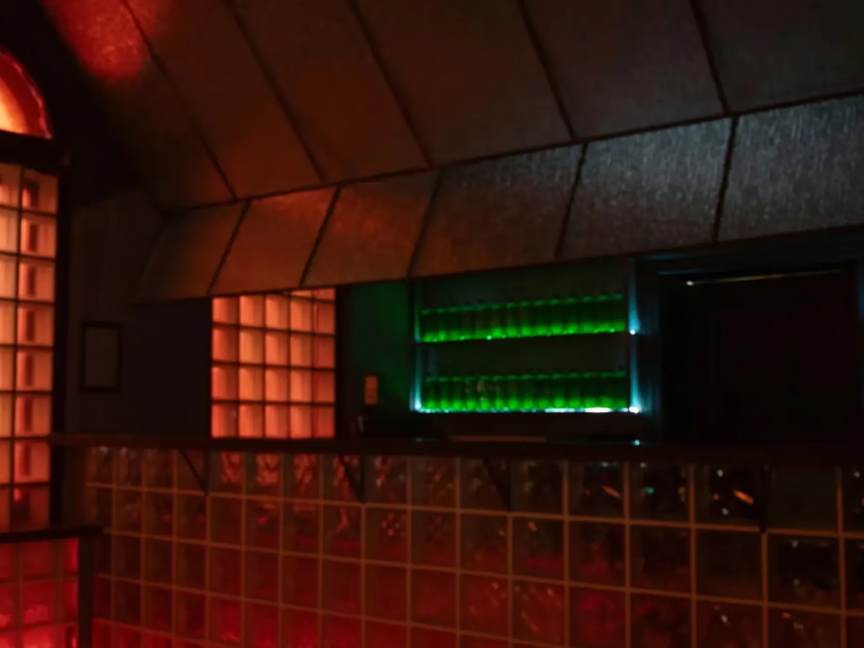 Bar mit grün beleuchteten Flaschen neben einem Glasbausteinfenster im Apartment