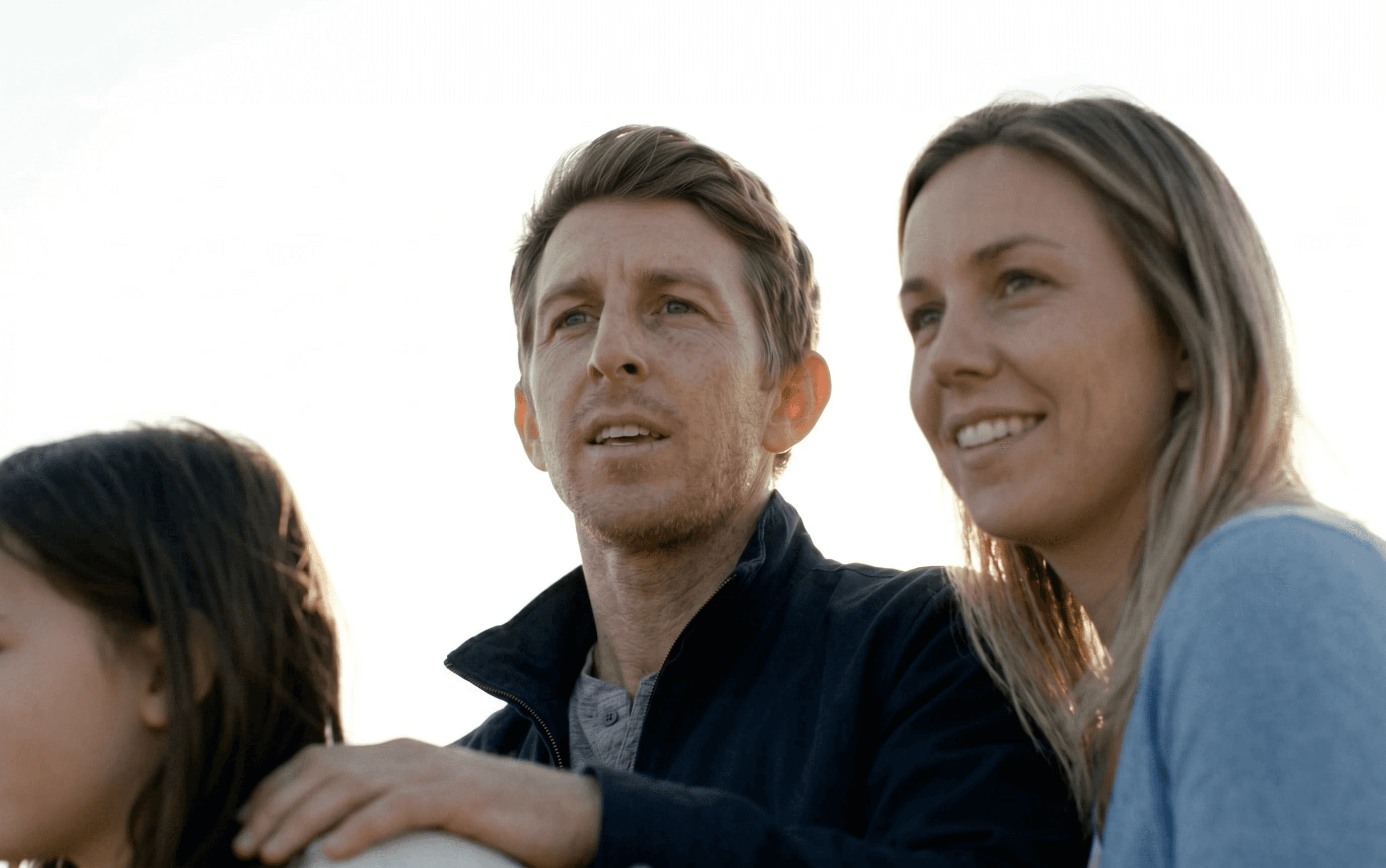 A family stands closely together outdoors under a bright sky, looking content and focused on something in the distance.
