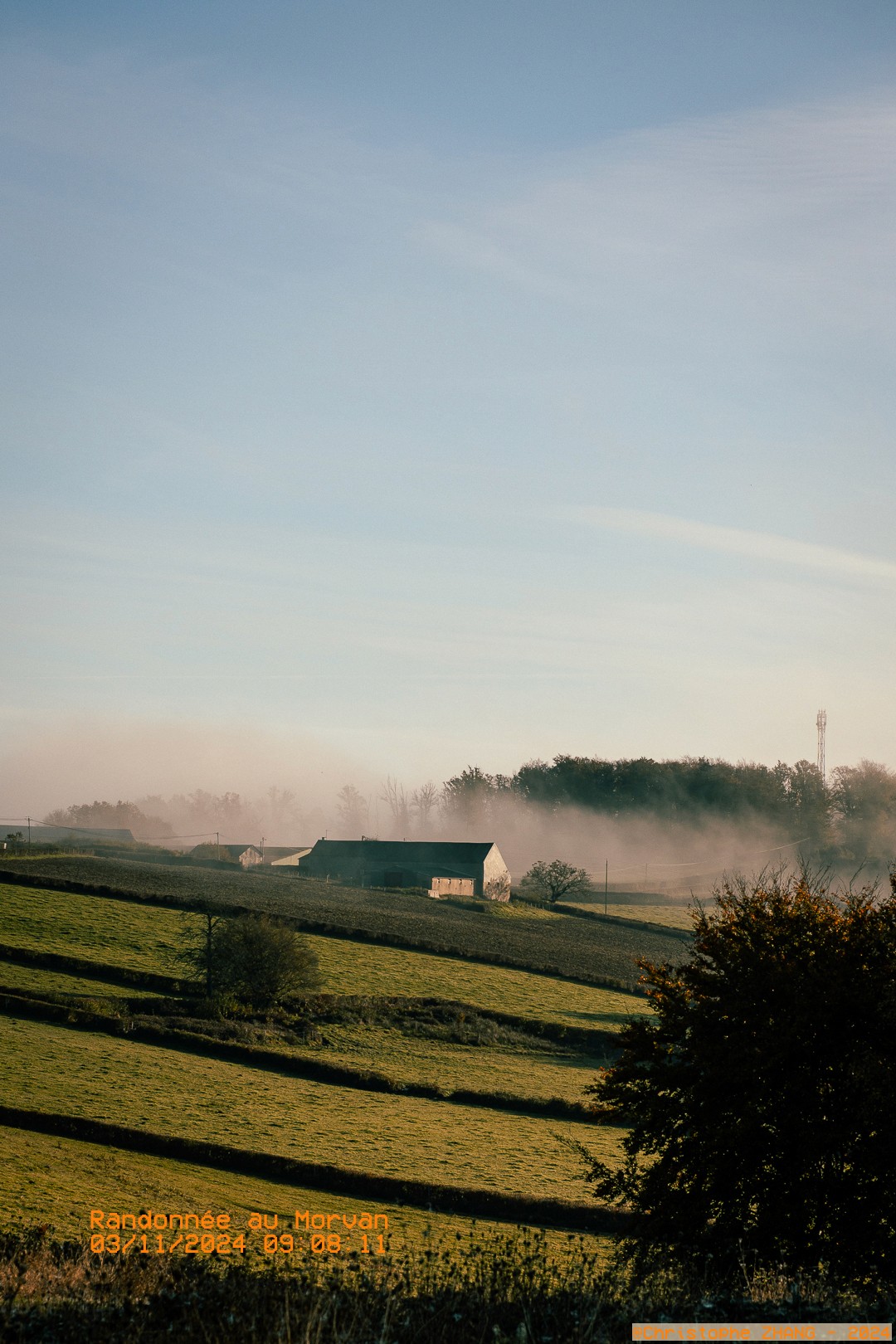 Paysage vallonné du Morvan au lever du jour, avec une ferme entourée de brume matinale.