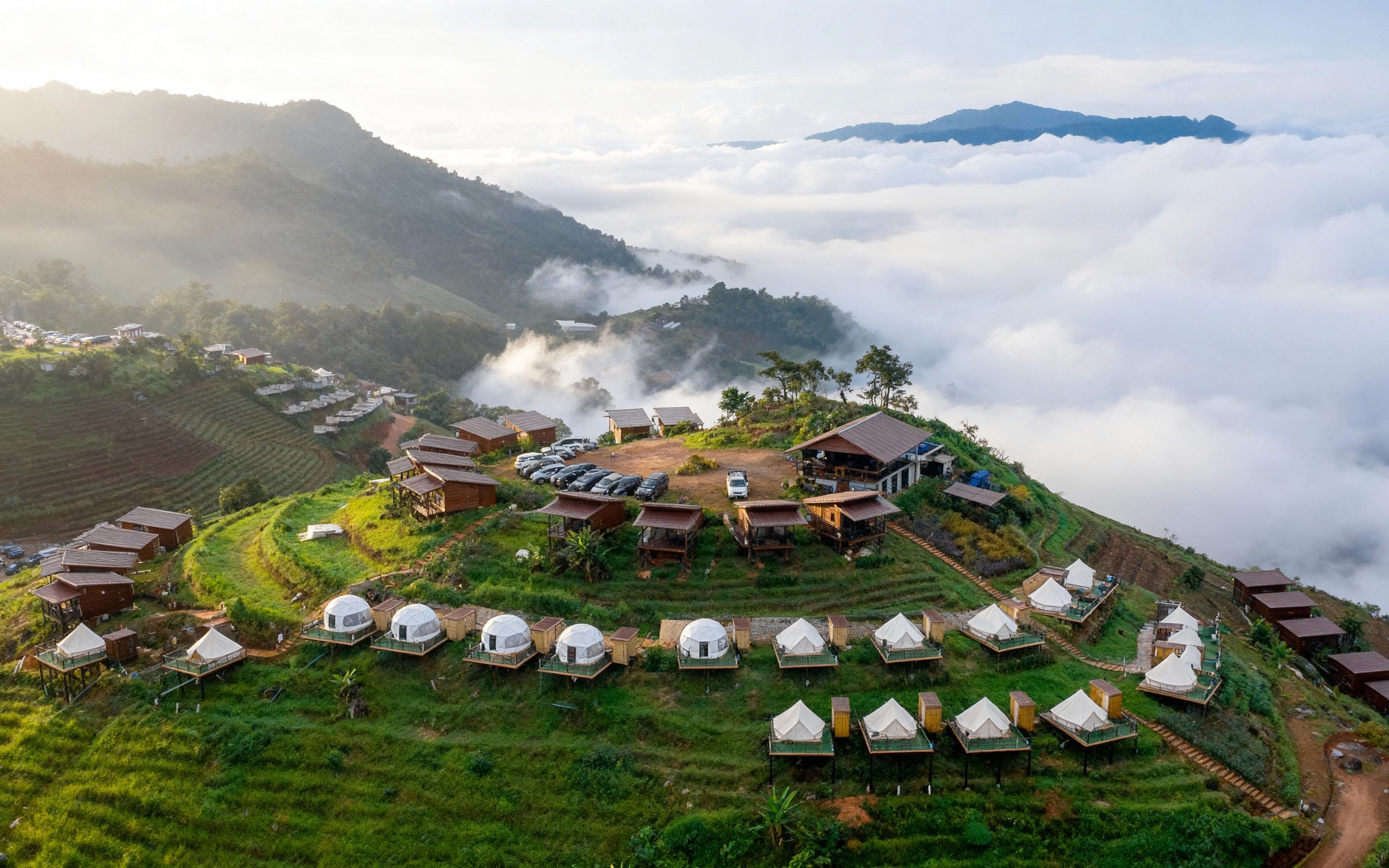 Aerial view of terraced hillside resorts and glamping tents surrounded by misty mountains and lush greenery in Mae Rim, Chiang Mai, with clouds rolling through the valley at sunrise.