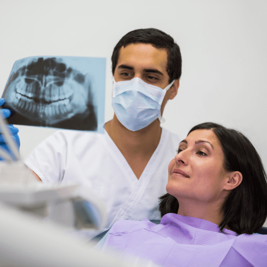 A dentist wearing a mask holding up a dental X-ray for a female patient in a dental chair, explaining the results.