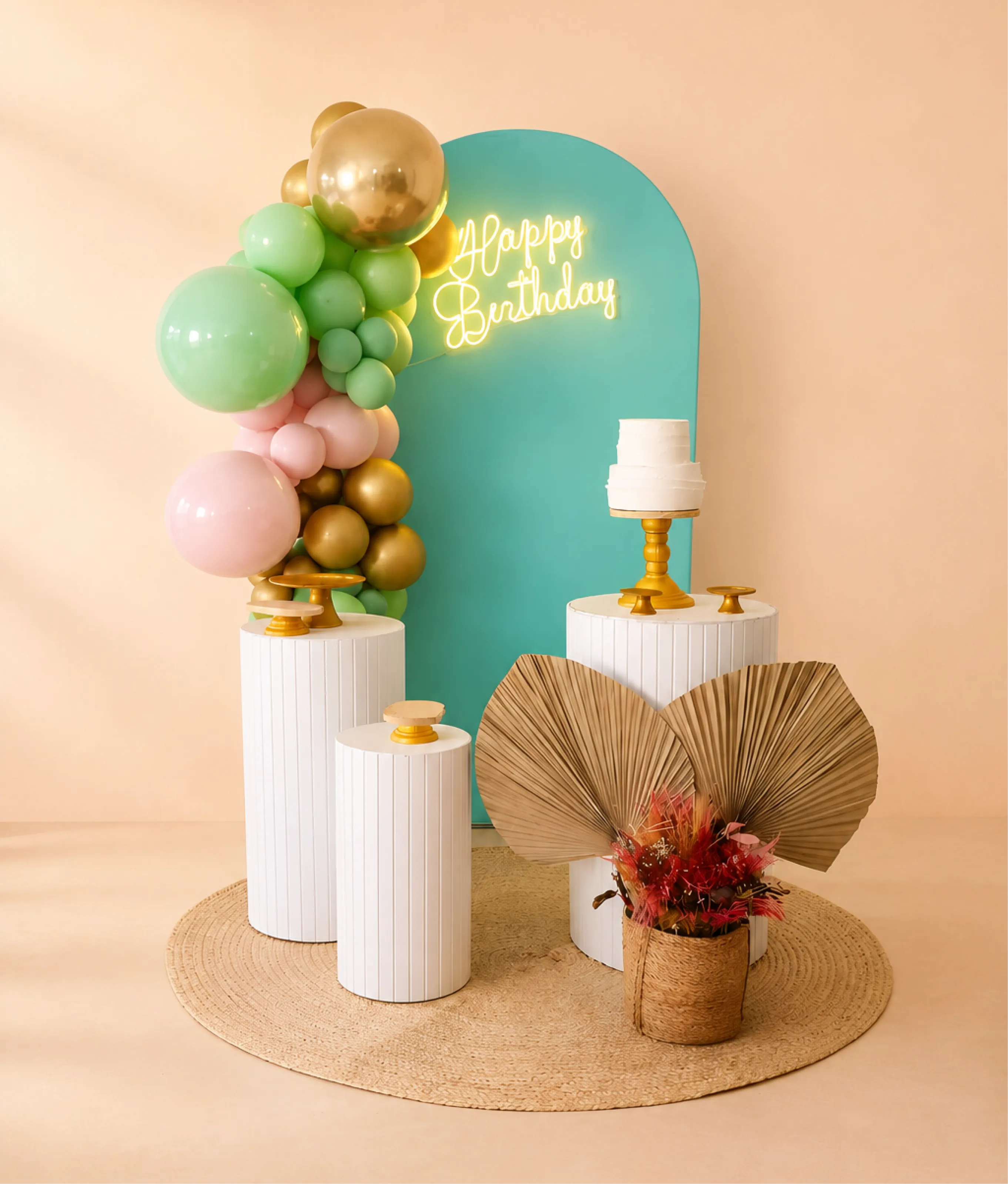 Decoração de aniversário com painel verde, arco de balões verdes, rosas e dourados, cilindros brancos, bolo e folhagens decorativas.