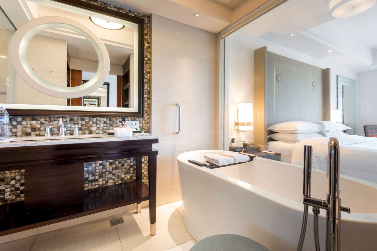 A modern bathroom features a large round backlit mirror over a dark wood vanity and a white soaking tub next to a bed.