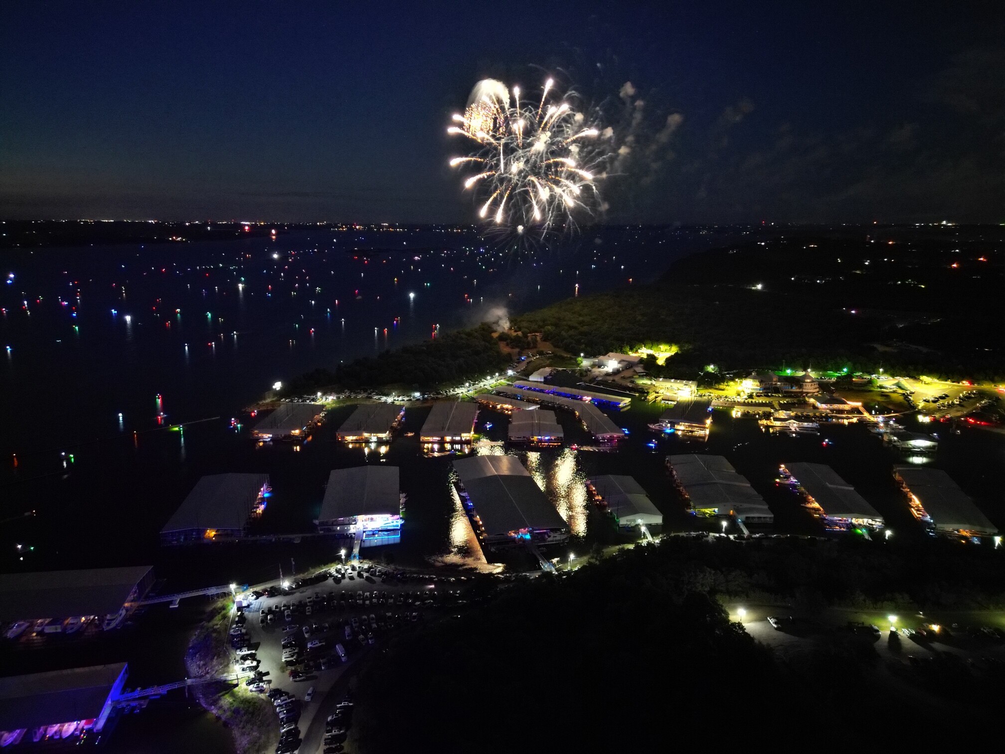 A vibrant fireworks display illuminates the night sky above a bustling waterfront marina, with colorful lights reflecting off the water and numerous boats dotting the bay.