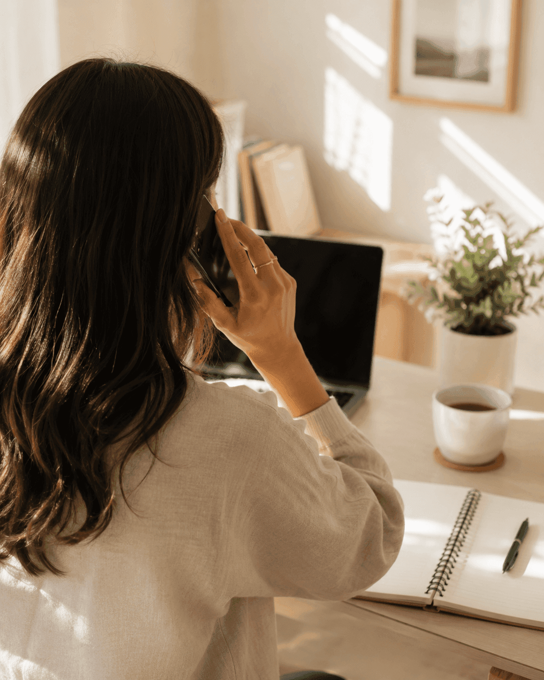 A woman speaking on the phone at a home desk with a notebook and coffee