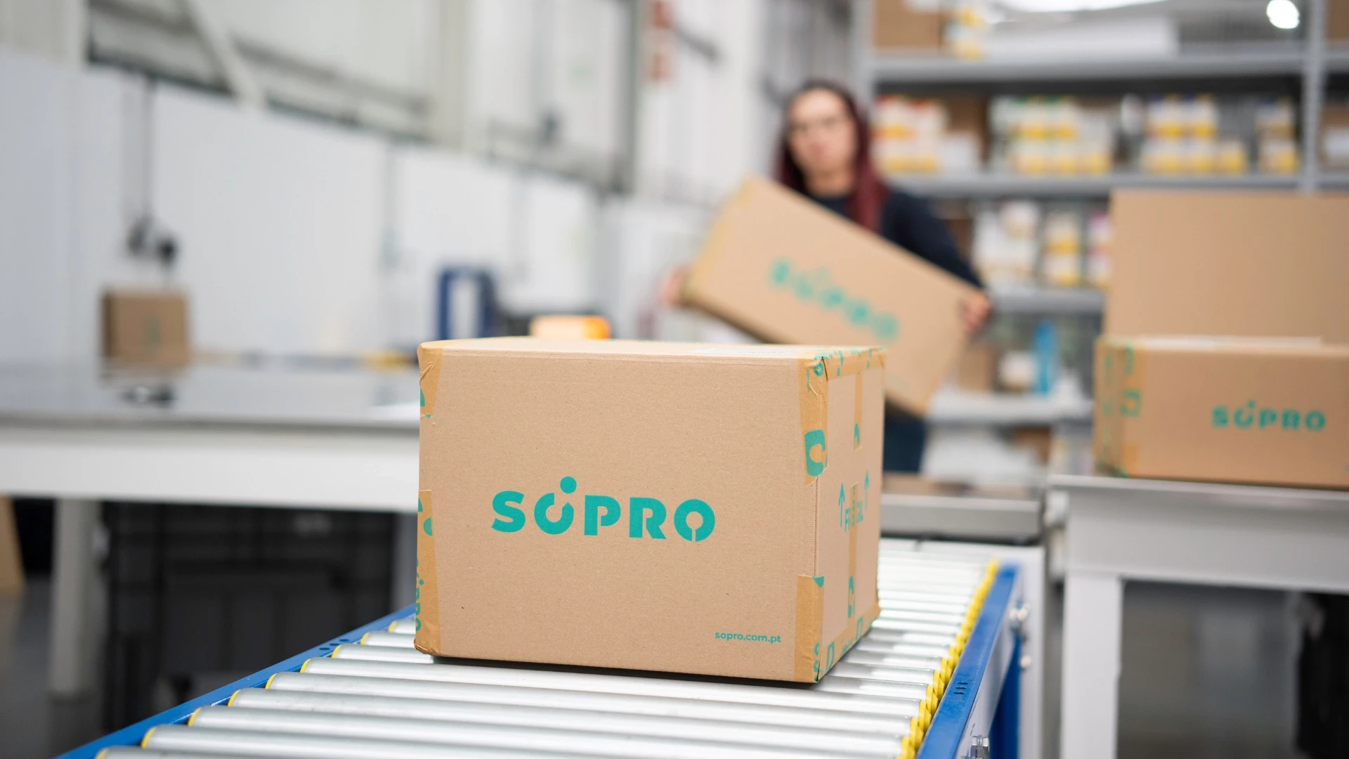 Imagem de uma embalagem com a marca Scipro em uma linha de produção, com uma pessoa segurando outra embalagem ao fundo, em um ambiente industrial.