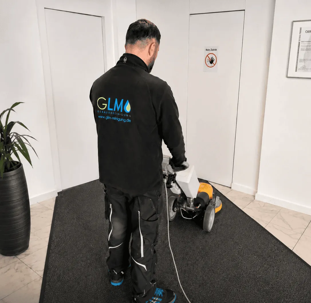 in Mann von hinten in dunkler Arbeitskleidung mit dem Logo 'GLM Gebäudereinigung' reinigt einen großen, dunklen Teppichläufer im Eingangsbereich mit einer gelb-weißen Einscheibenmaschine