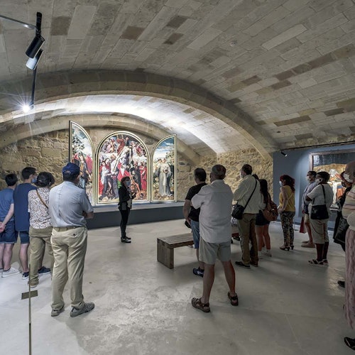 A group of people observes a large triptych painting in a stone-arched gallery room, with a guide providing information.