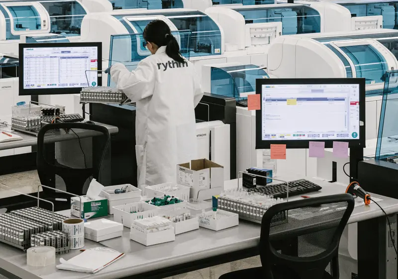 Laboratory technician working with automated testing machines and sample trays in a clinical lab environment.