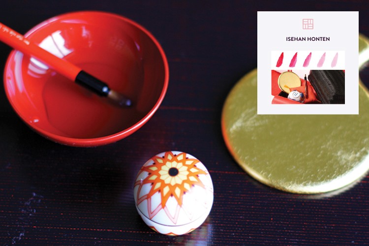 An orange bowl containing a paintbrush rests on a dark wooden surface. Next to it, a small white ball with orange and yellow floral patterns sits on the surface. A gold circular object is positioned to the right of the ball. In the top right corner, a white card displays a logo and the text "ISEHAN HONTEN" above three small images of lipstick swatches.