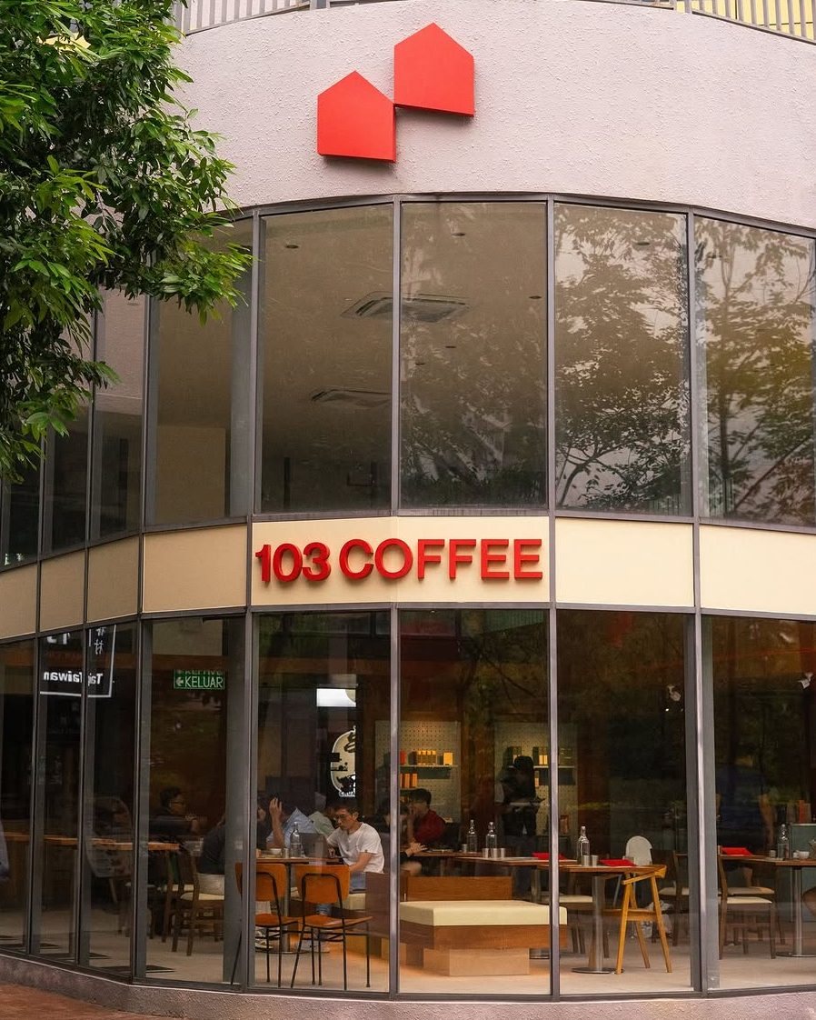 Exterior view of 103 Coffee café with large glass windows, showcasing seating area and vibrant red signage.