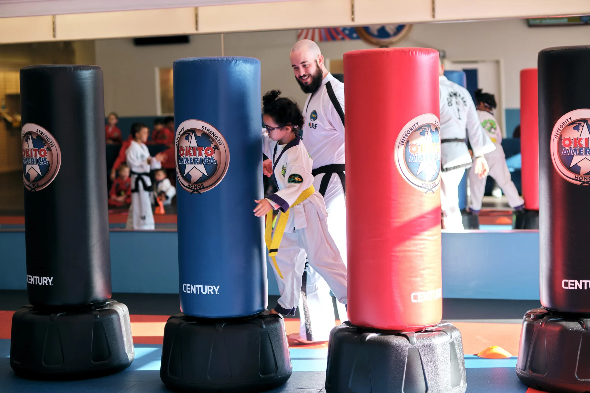 An instructor guides a young student in a yellow belt through a martial arts technique while standing between colorful Century brand heavy bags in a dojo training facility.