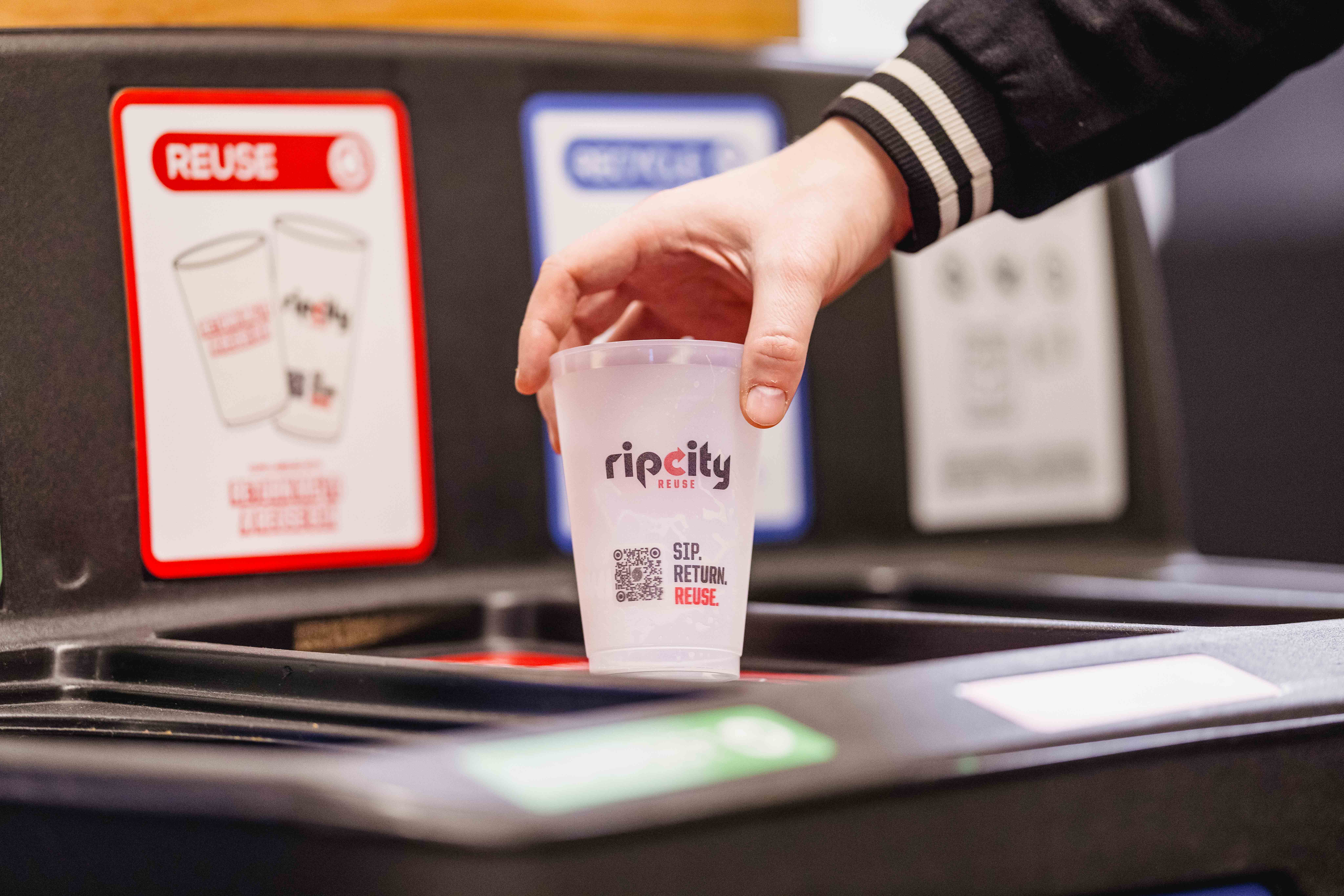 Hand placing an empty Rip City Reuse cup into a Moda Center reuse return bin, with signage for reuse and recycling stations in the background.