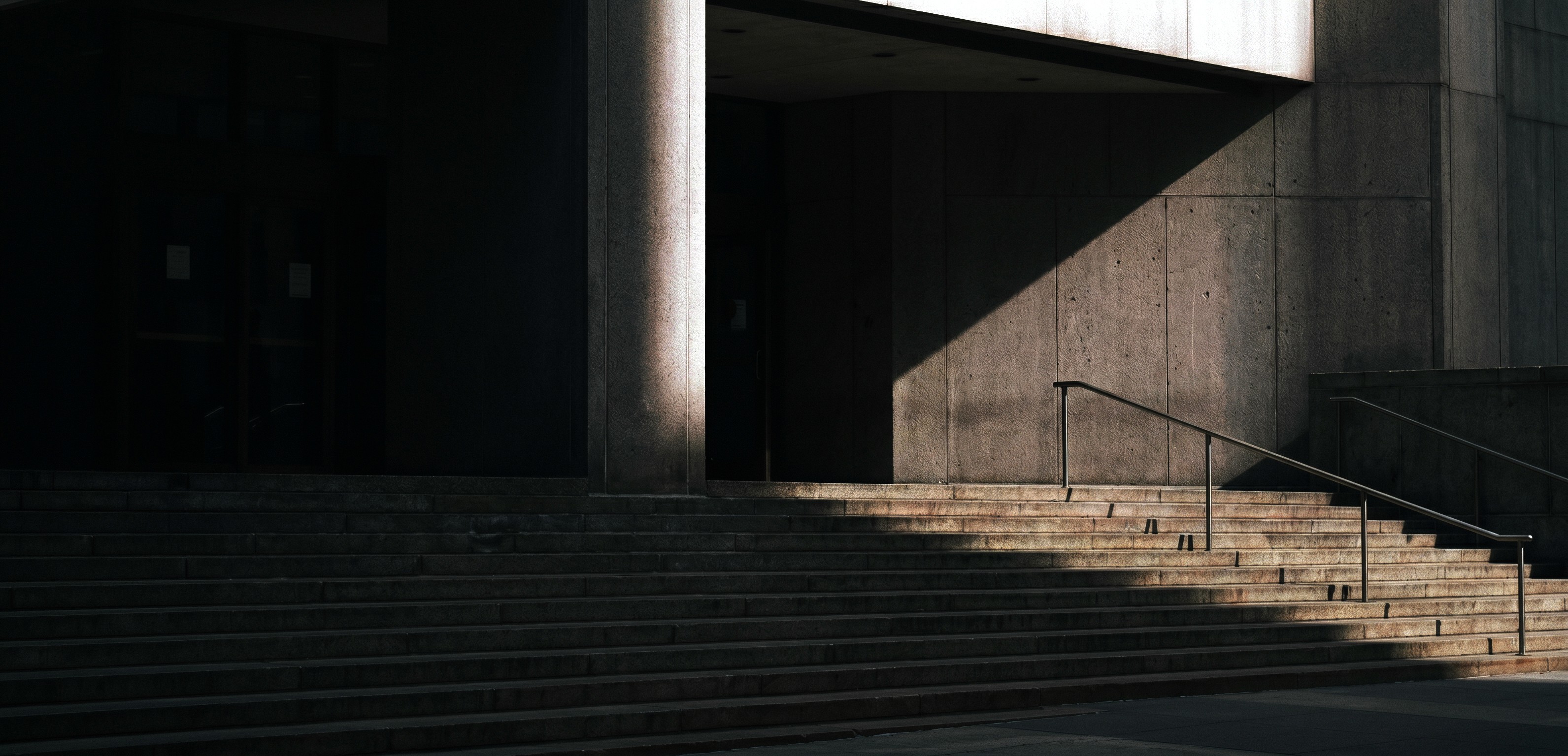 Wide stone staircase leading to a shadowed building entrance, featuring a metal handrail and dramatic sunlight contrast, creates a sense of urban architecture and design.