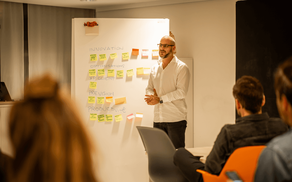 Homme en chemise blanche qui présente devant un tableau avec des post-it colorés face à une audience assise l’écoutant.