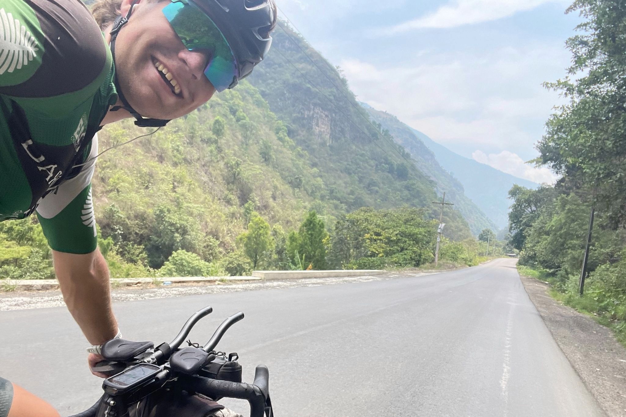 A cyclist in a helmet and sunglasses take a selfie while he cycles along an empty road in Central America with lush hillsides on each side