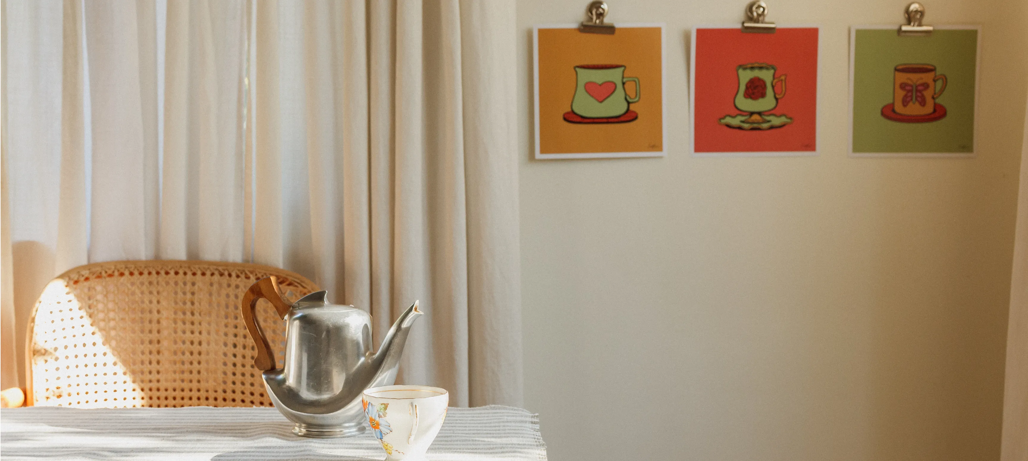 A distant shot of a dining table with a linen table cloth, a silver tea pot, a tea cup, and 3 prints hanging on the wall behind. 