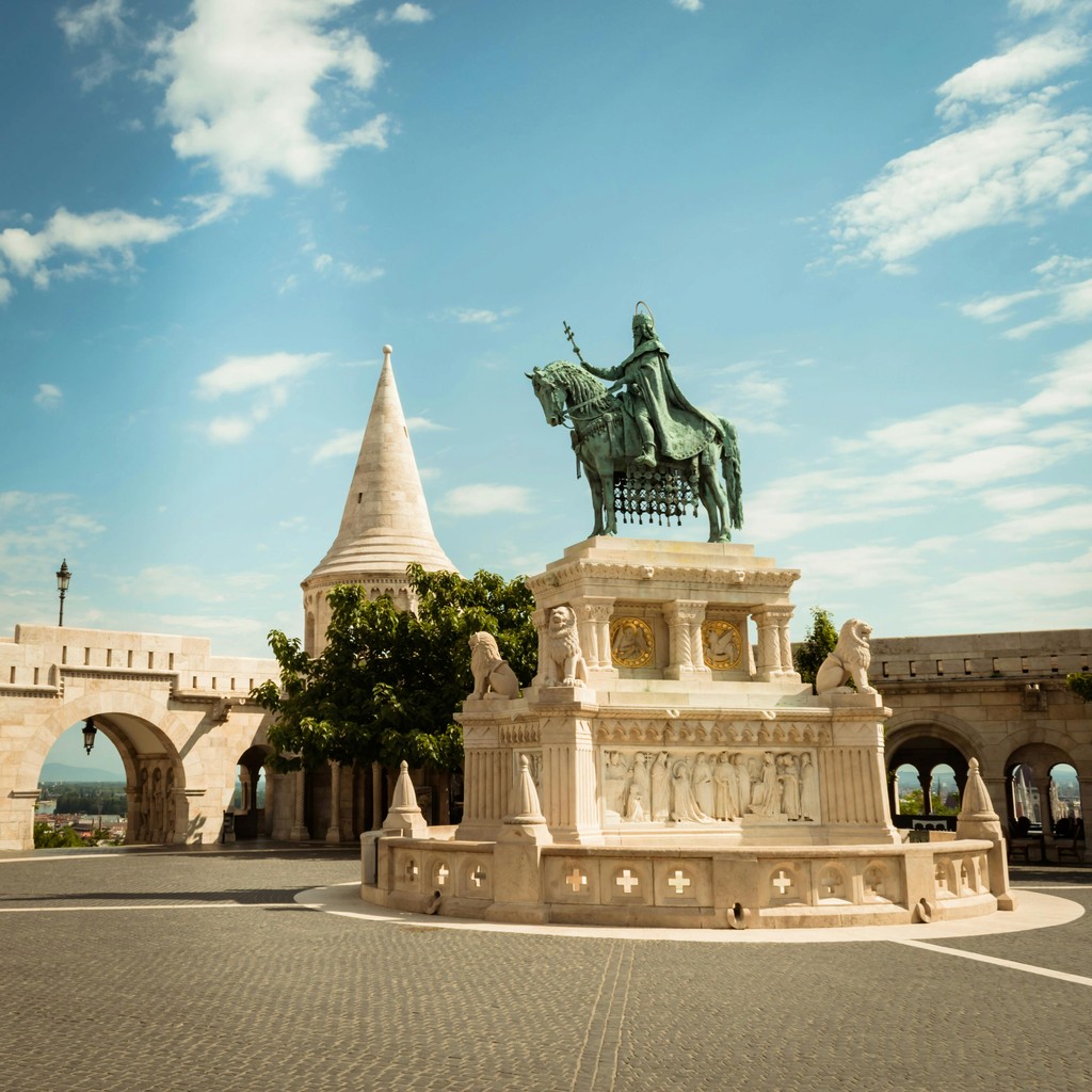 The image showcases the iconic statue of Stephen I of Hungary, mounted on a horse, located in Fisherman's Bastion, Budapest. The statue is set against a backdrop of historical architecture, including pointed towers and arched colonnades, under a bright, clear sky. This landmark is a significant tourist attraction, symbolizing Hungary's rich history and architectural heritage.