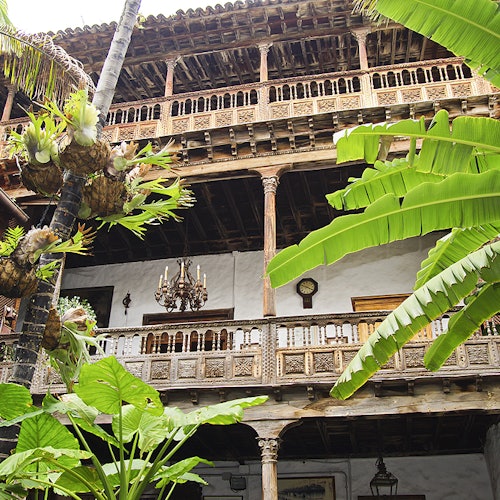 Three-story building with ornate wooden balconies and lush tropical plants in the foreground. A chandelier hangs on the second floor.