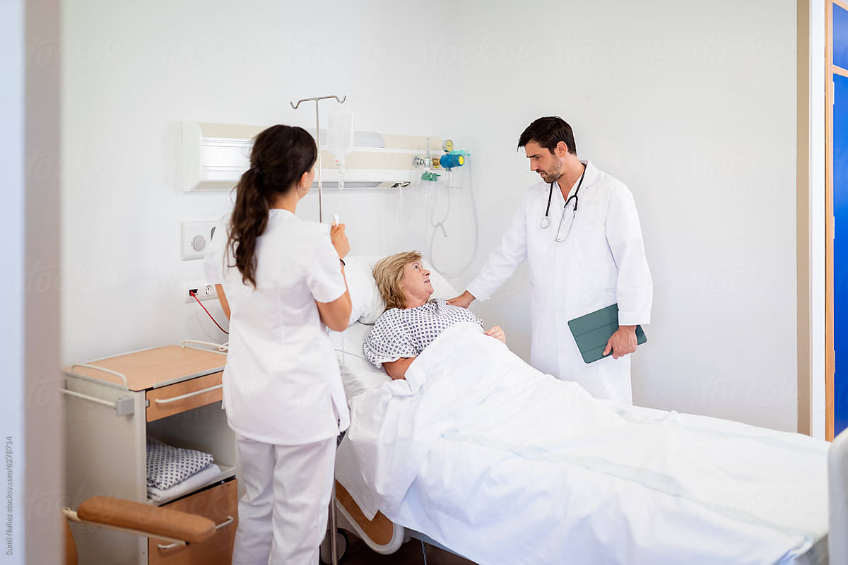 A healthcare professional in a mask and scrub uniform providing compassionate care to an elderly patient in a hospital bed. 
