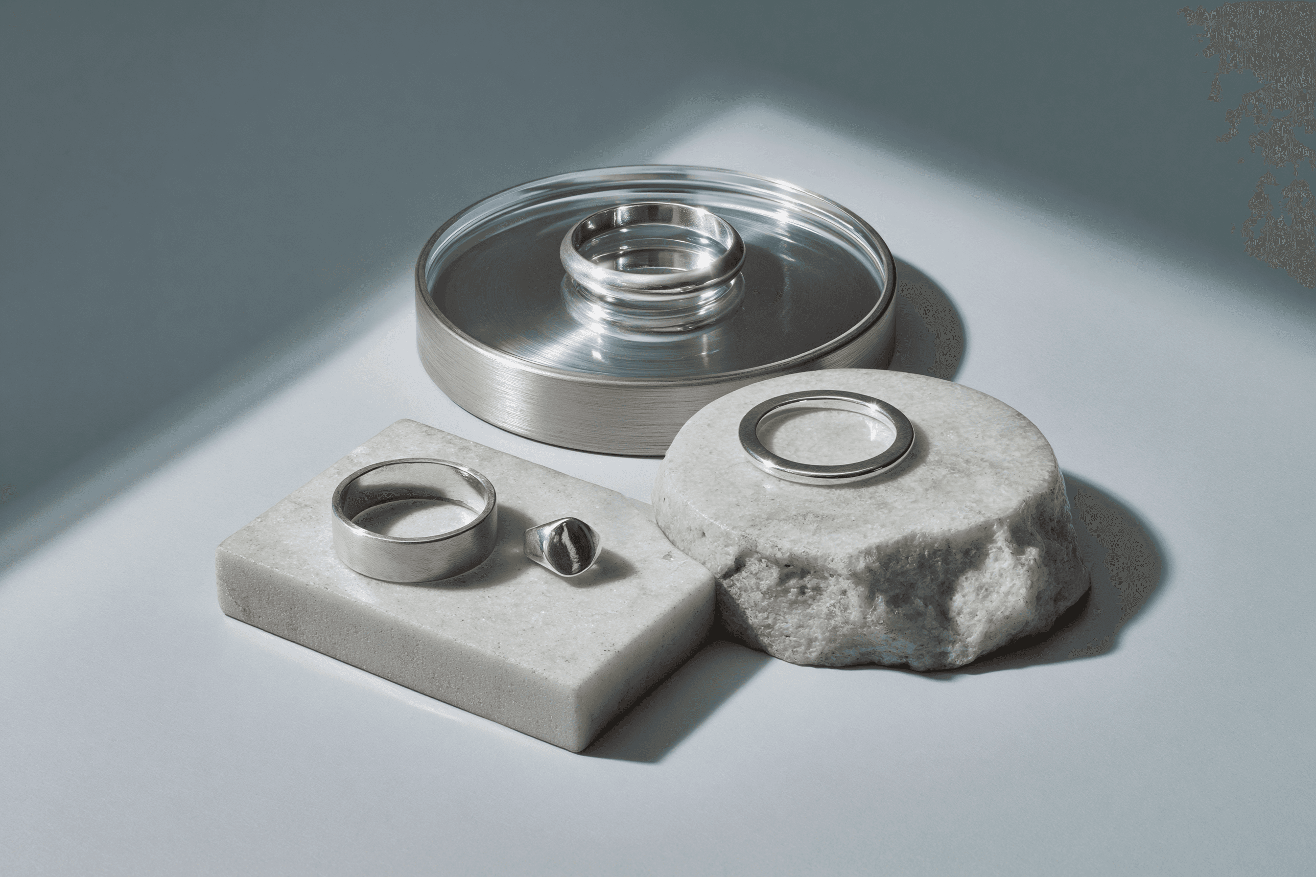 A collection of silver rings displayed on white stone pedestals and a reflective glass surface, with soft shadows casting across a light gray background.