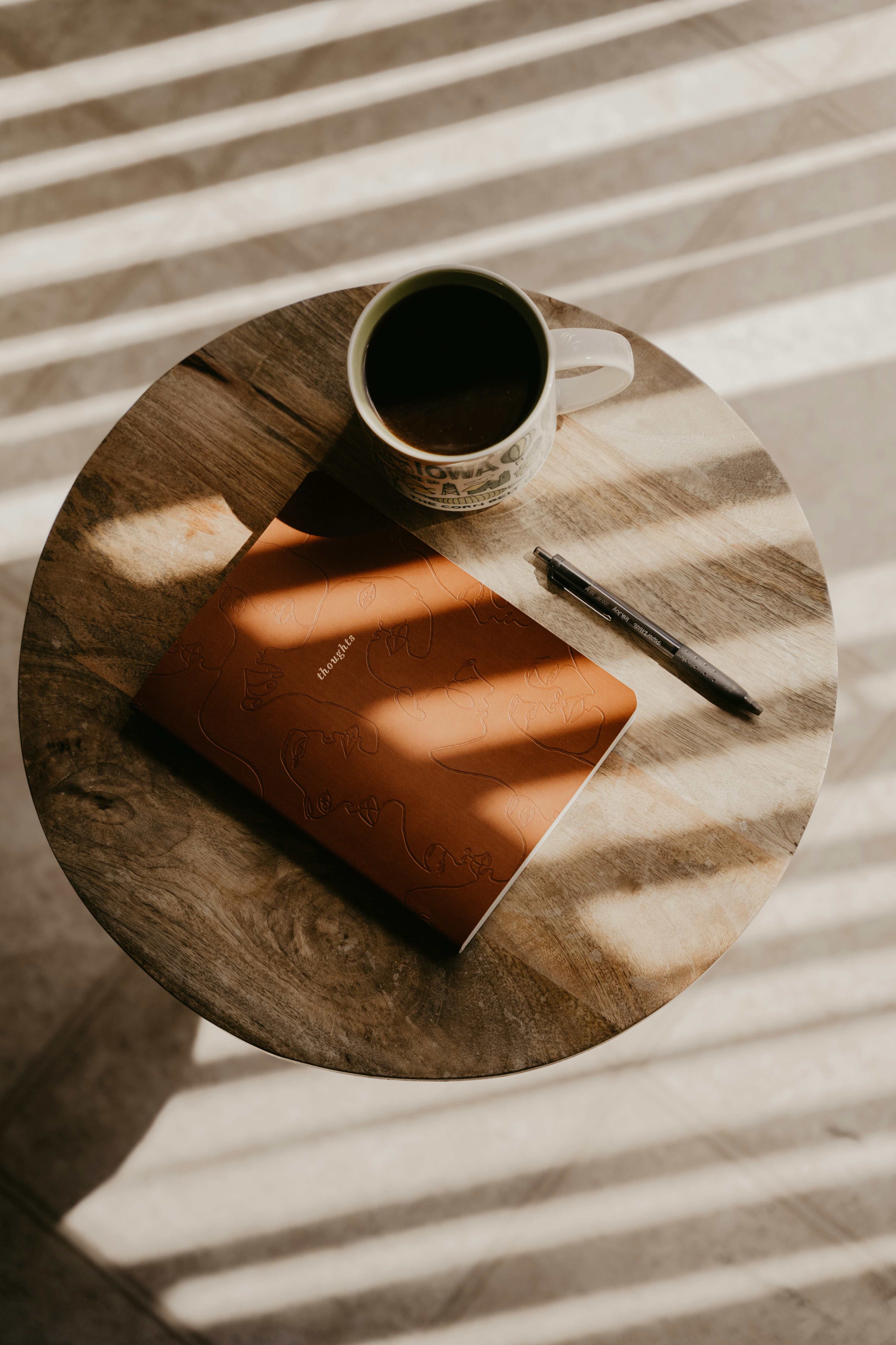 Notebook and pen on a round wooden table beside a cup of coffee, lit by soft sunlight through blinds.