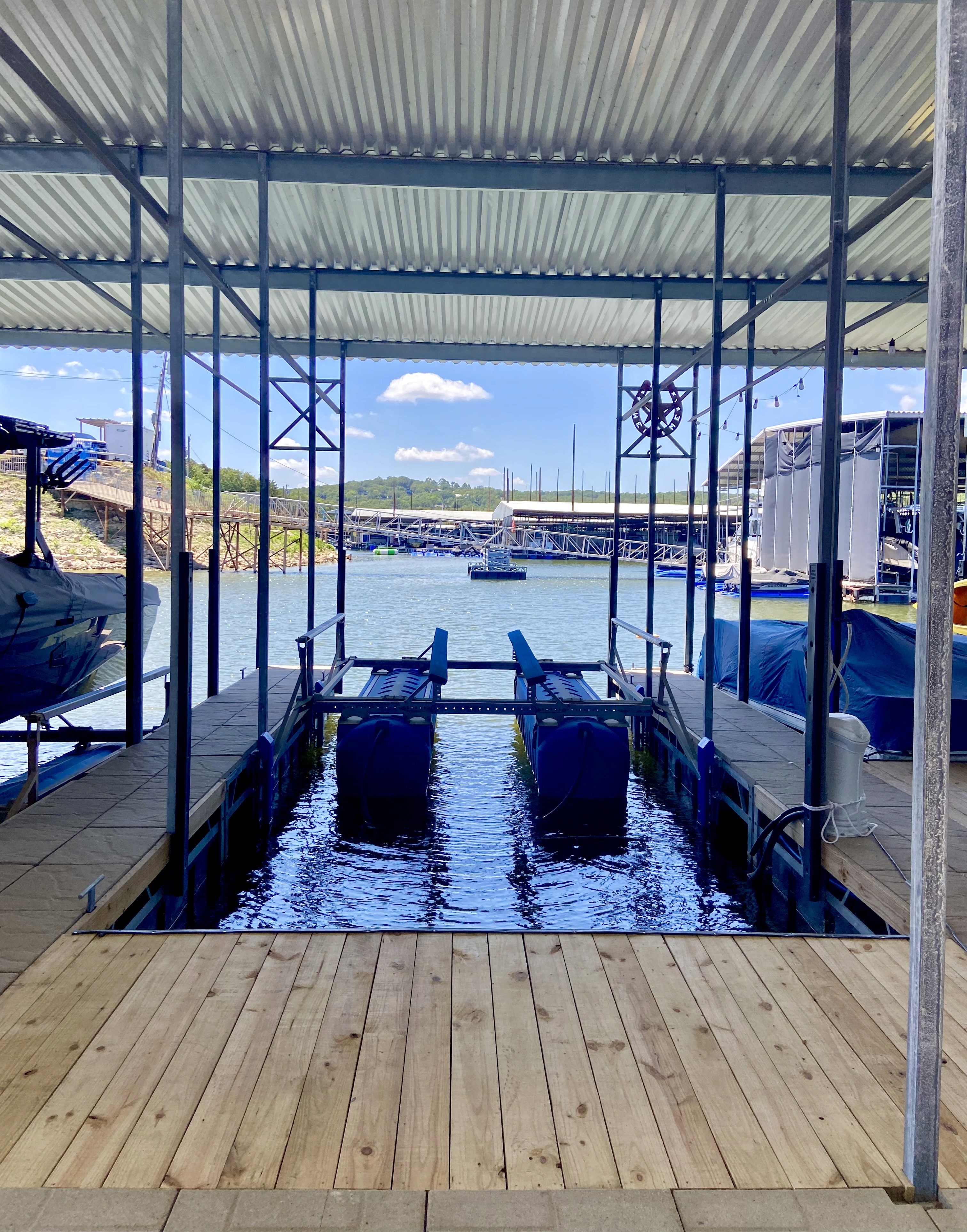 A covered boat lift at a waterfront marina with blue steel beams and a wooden dock in the foreground, overlooks a serene lake framed by green hills under a clear blue sky with a few scattered clouds.