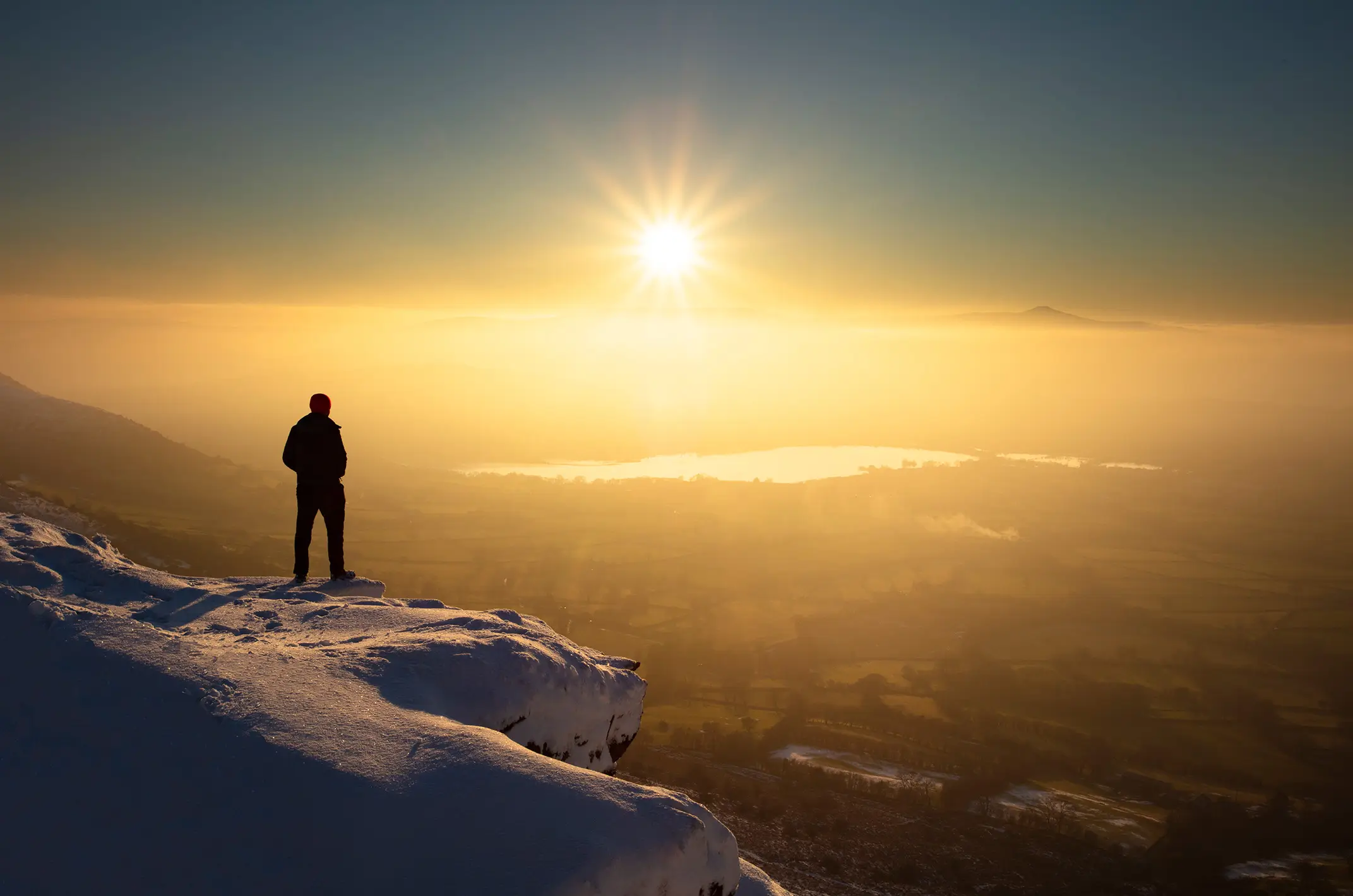 A lone figure stands on a snowy peak, gazing at a vibrant sunset illuminating the sky.
