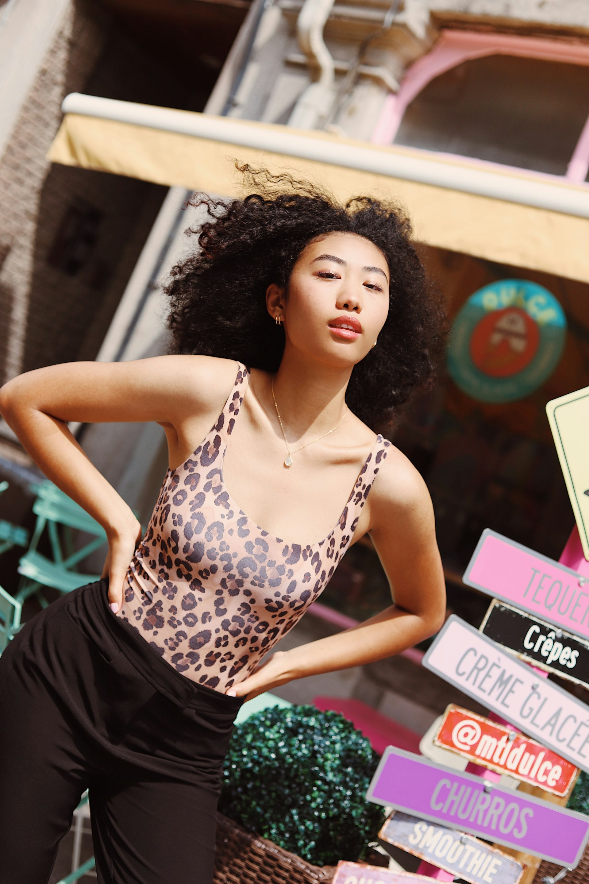 A woman with curly hair poses outdoors near colorful shop signs. She is wearing a leopard-print bodysuit and black trousers, looking directly at the camera with a neutral expression.