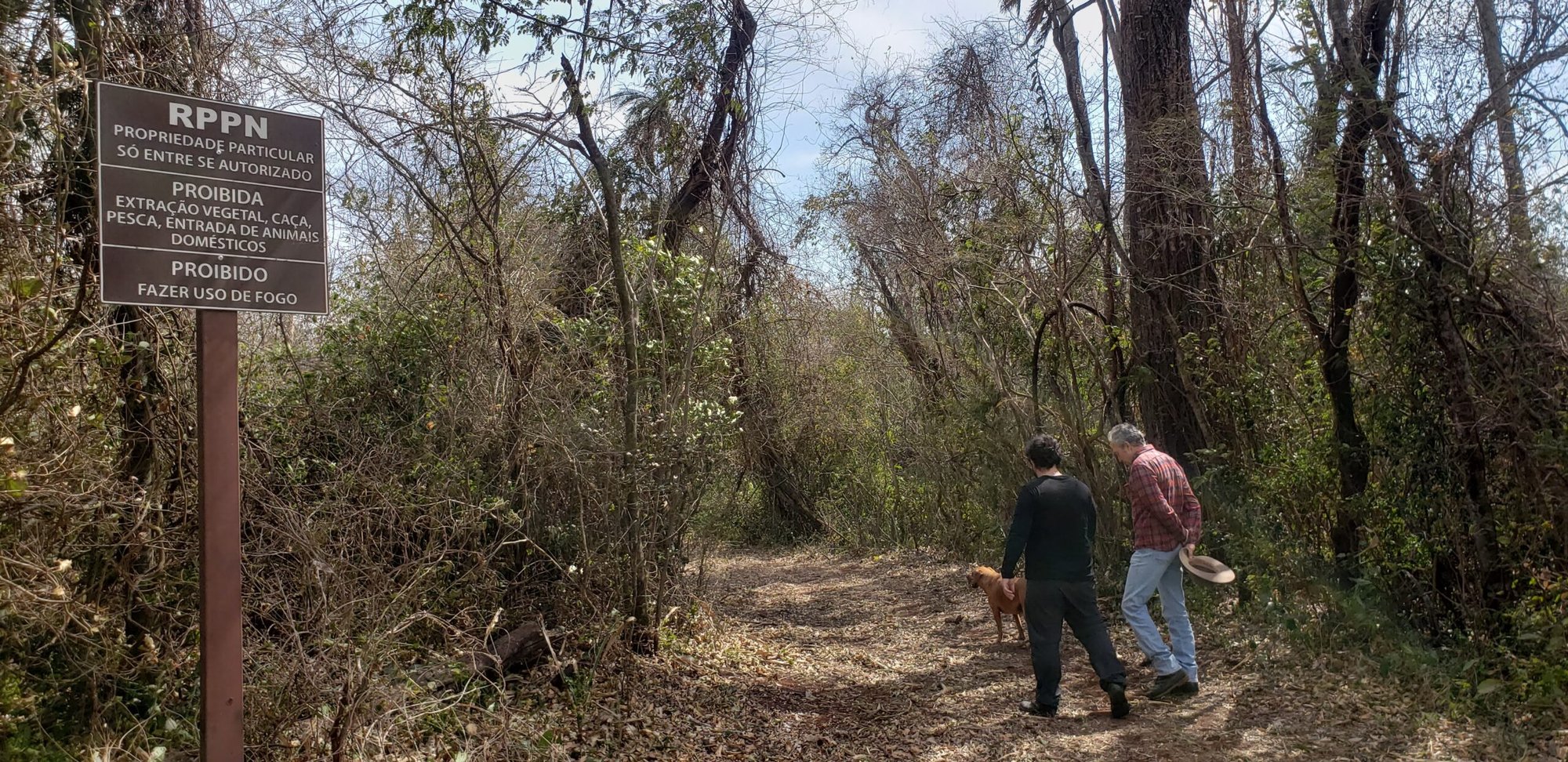 Duas pessoas e um cachorro caminham em trilha de mata seca ao lado de placa da RPPN com proibições