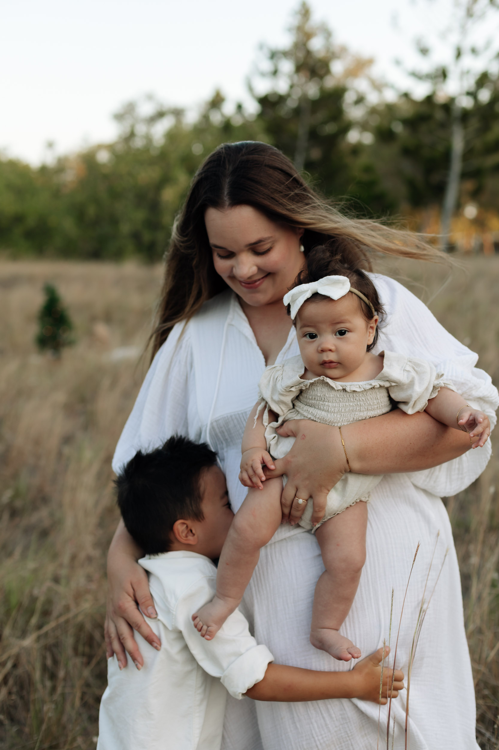 Sunset family session in tall grass in Mackay with mother holding baby girl and young boy standing cuddling