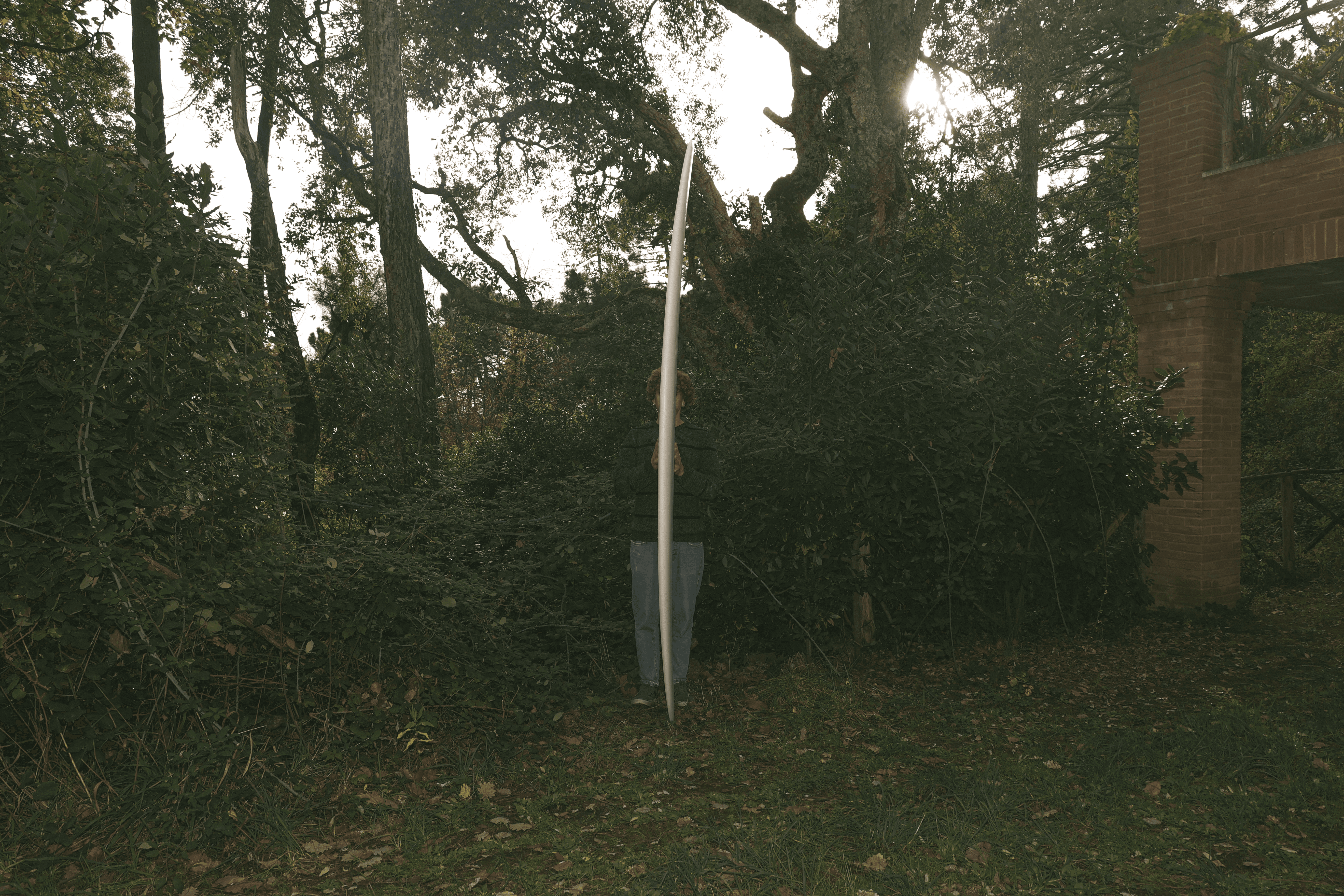 Una persona in piedi in un boschetto verde, tiene in verticale una longboard da surf bianca e stretta che nasconde in gran parte il suo corpo.