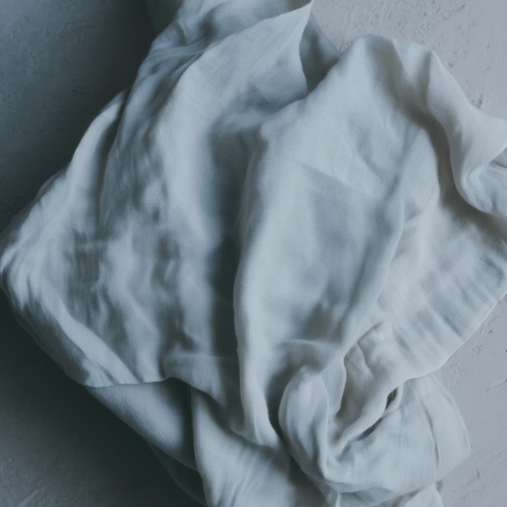 A crumpled, soft white fabric lies against a textured gray surface, showcasing gentle folds and subtle shadows that highlight its delicate, airy quality.