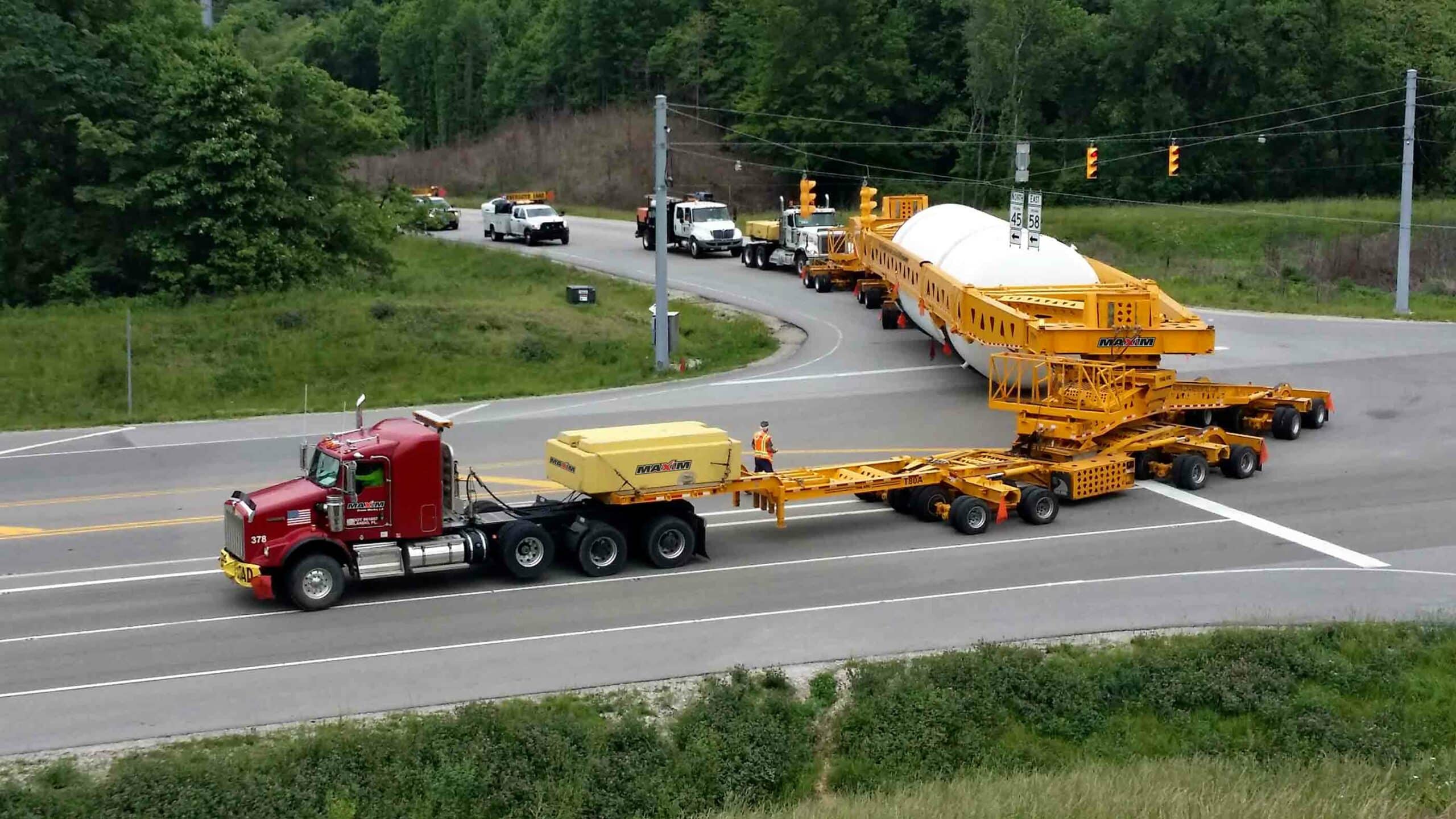 A heavy flatbed red-cab truck with dozens of axles, carrying a large white cylinder, making a turn to enter a road accompanied by several other small trucks and a guy in yellow helping with the maneuver.