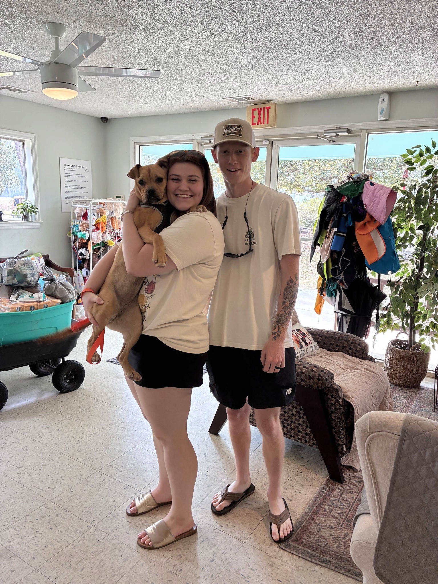 A couple stands together in a bright room, holding a small dog and a child, all smiling.