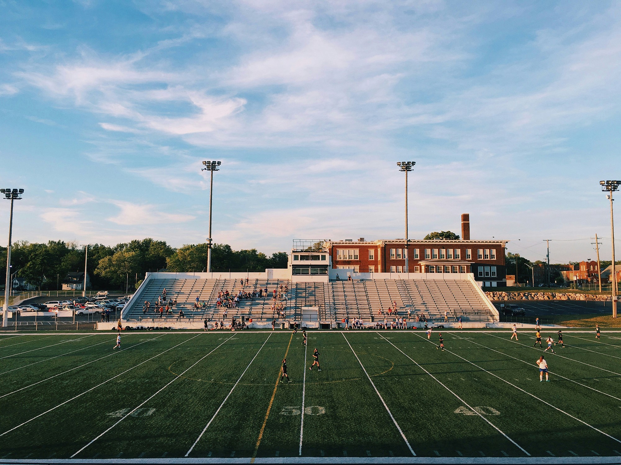 A sports field at sunset, featuring stands and floodlights against a blue sky with scattered clouds.