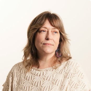 Portrait of a woman with medium-length wavy light brown hair with bangs, wearing a cream crocheted top and colorful drop earrings, looking slightly to the side against a plain white background.