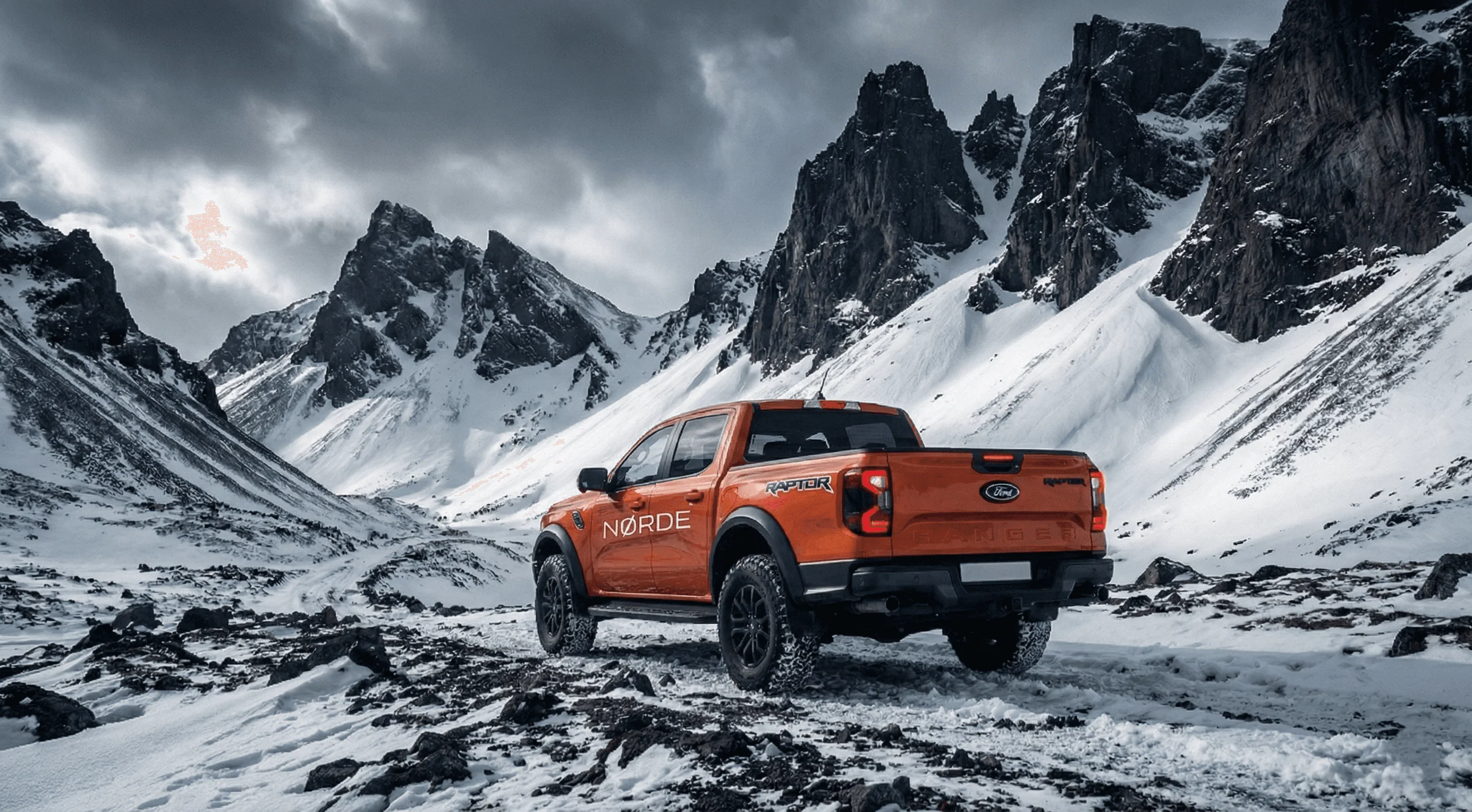 An orange Ford Ranger Raptor parked on snow in front of a steep, snow-covered mountain slope.